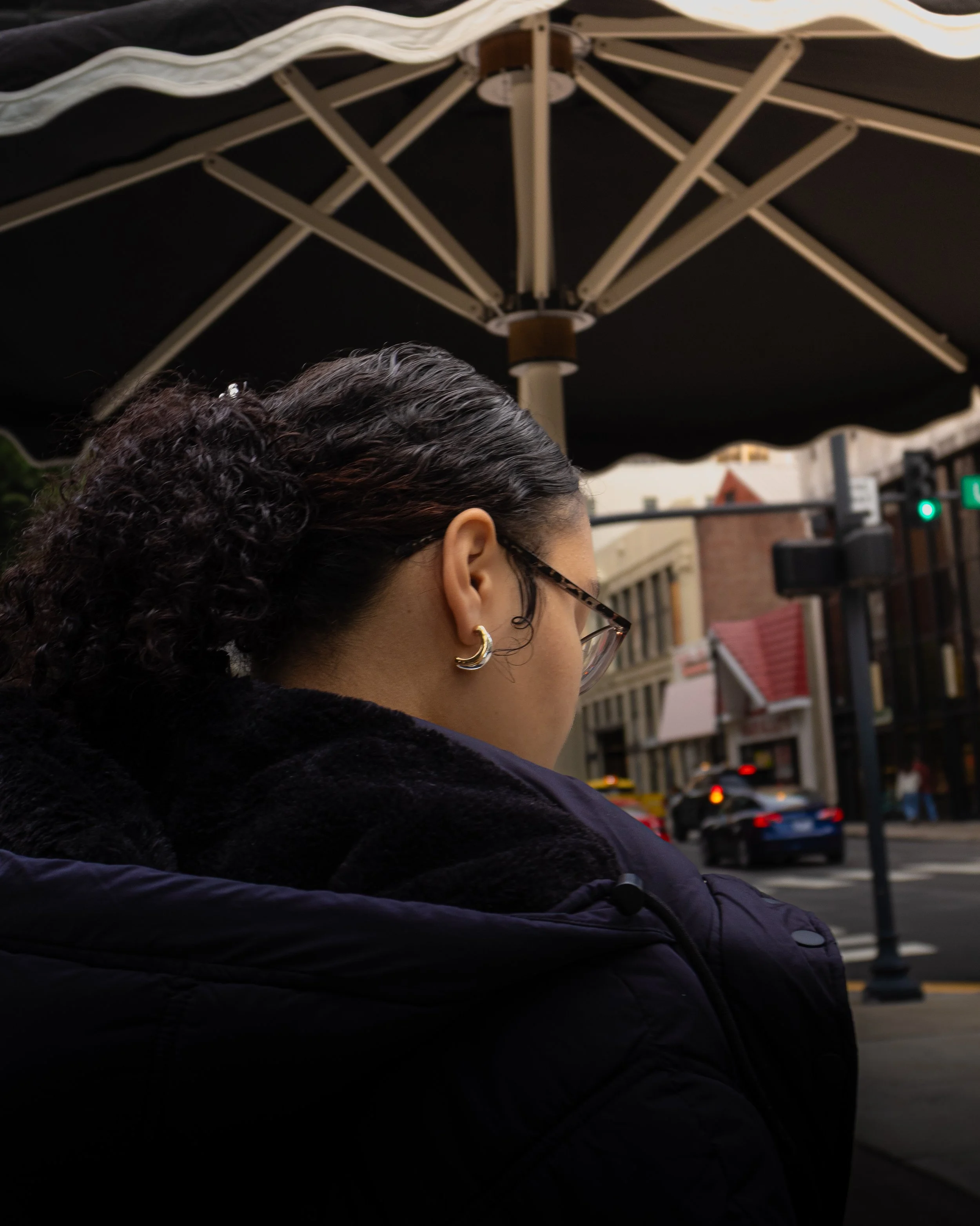 Close-up of a woman with curly dark hair, wearing glasses, earrings, and a dark jacket, standing outdoors near a city street with buildings, traffic lights, and cars in the background, under a large patio umbrella.