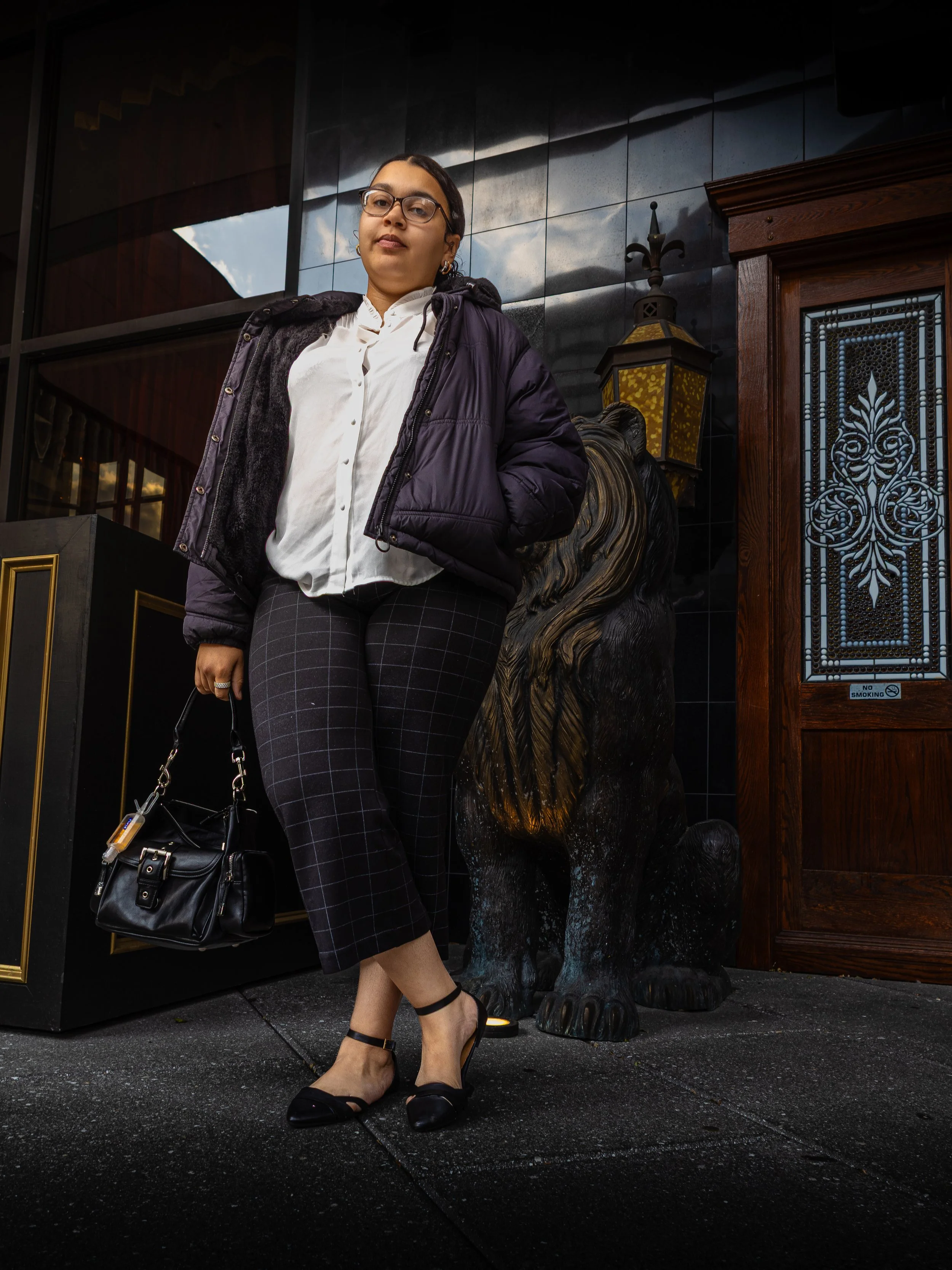 A woman standing outdoors near a lion sculpture. She is wearing glasses, a white blouse, a dark puffy jacket, plaid pants, and black heels. She is holding a black handbag.
