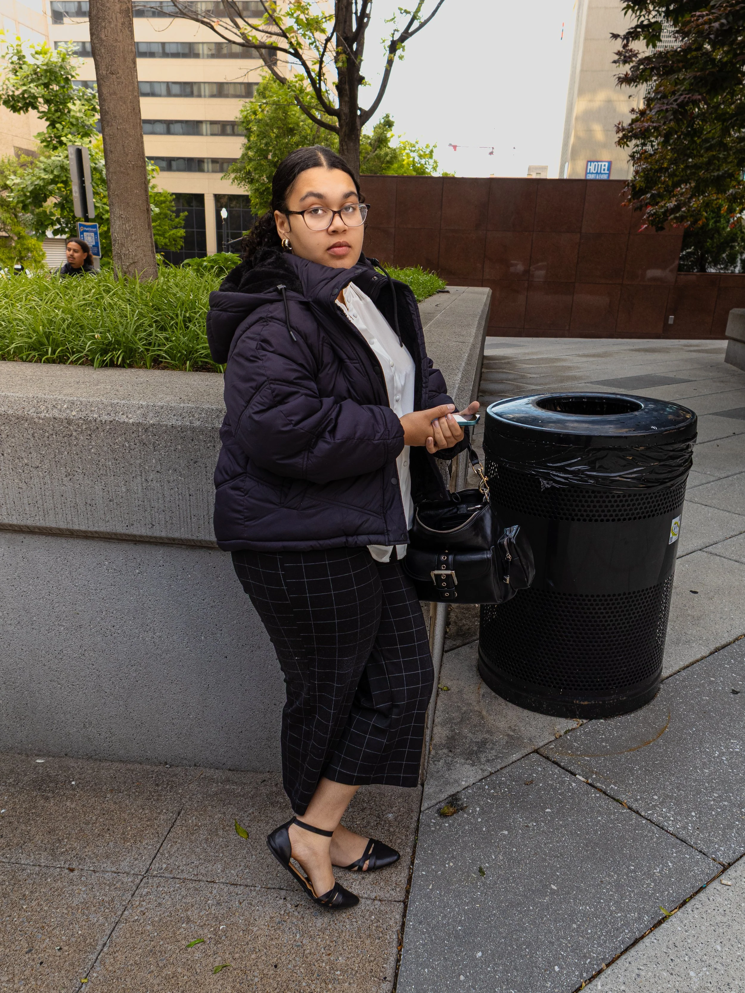 A young woman with glasses standing on a city sidewalk, holding a smartphone, wearing a dark jacket, white shirt, checkered skirt, and black shoes, near a black trash can, with trees and tall buildings in the background.