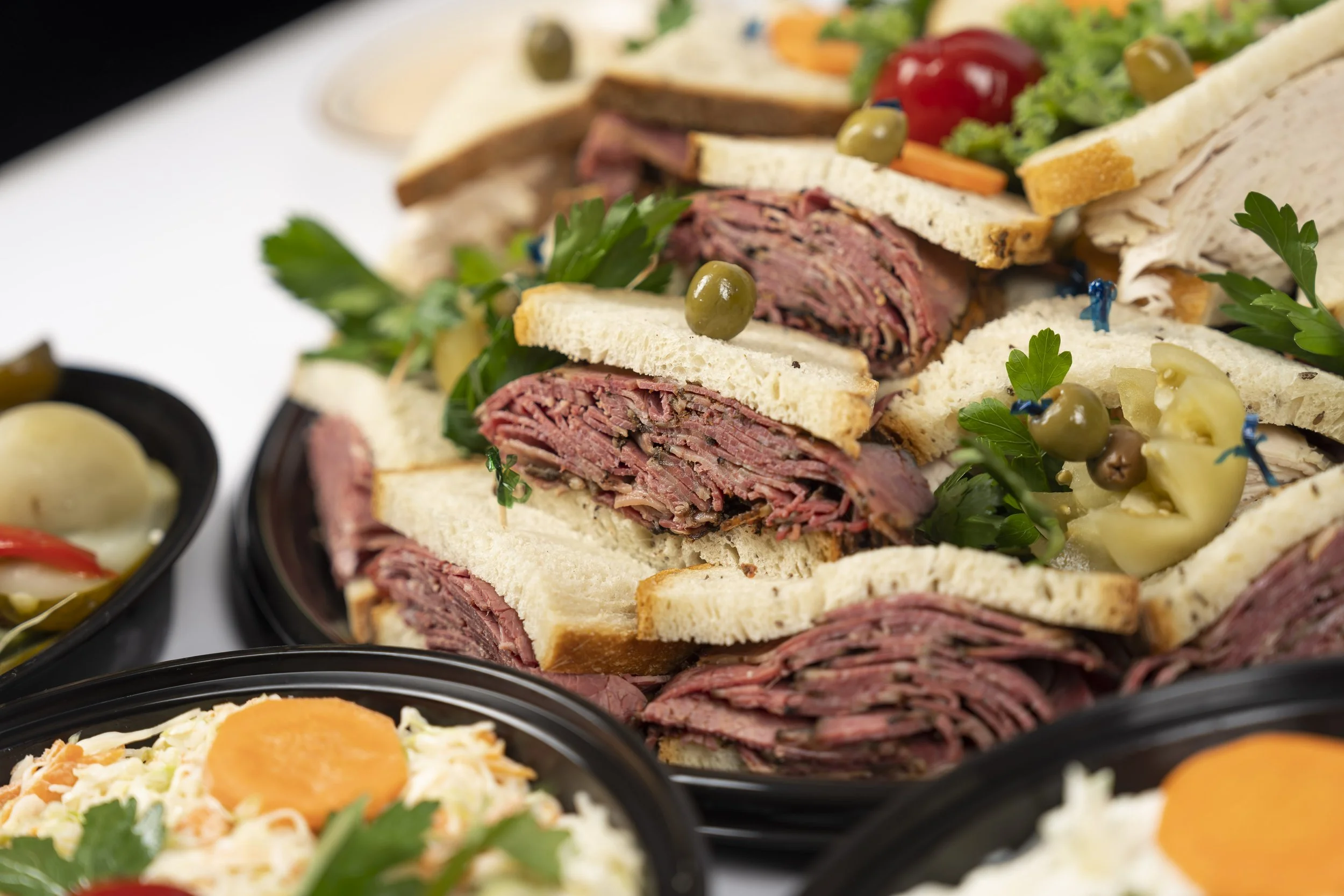 Near a stack of roast beef and turkey sandwiches with lettuce and other toppings, there are bowls of potato salad with carrots and boiled egg, and pickled vegetables in black bowls.