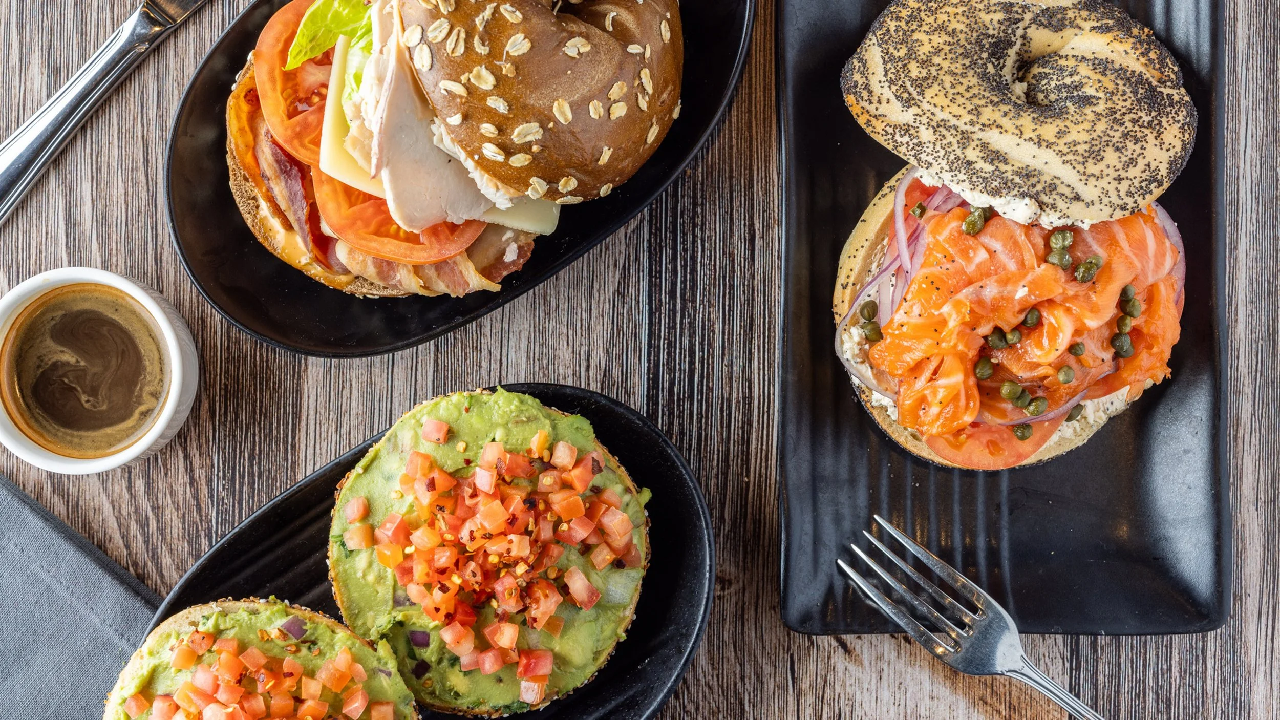 Assorted open-faced sandwiches and bagels with toppings on black plates, a cup of coffee, and a gray napkin on a wooden table.