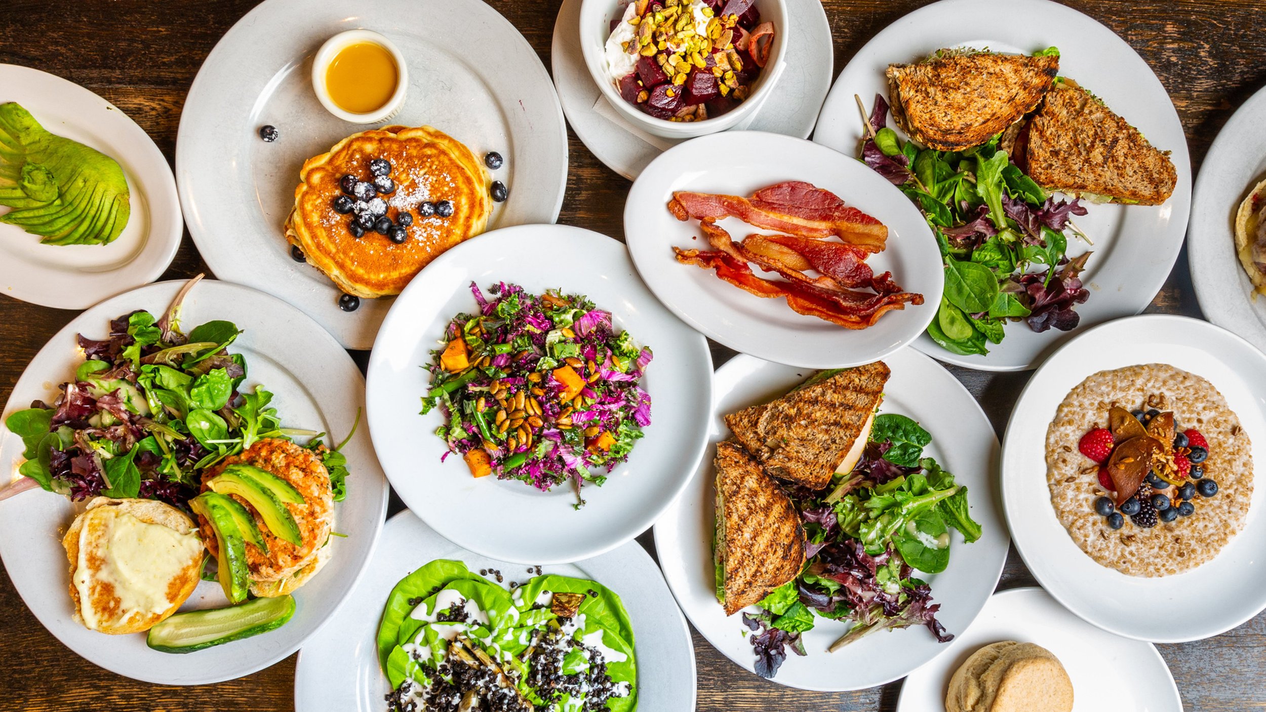 Assorted breakfast dishes including pancakes with blueberries, bacon, grilled sandwiches, salads with greens and vegetables, a bowl of oatmeal topped with berries and nuts, and a small bowl of syrup, all on white plates on a wooden table.