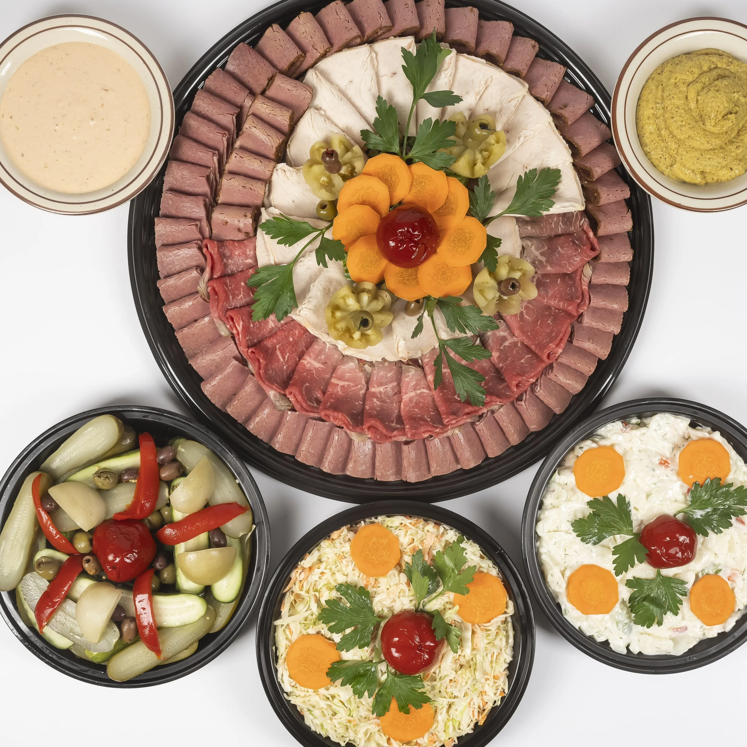 Assorted cold cuts, including slices of ham, roast beef, and salami, arranged in a circular pattern on a tray, topped with garnishes of parsley, carrot slices, and a cherry tomato. Surrounding bowls contain dipping sauces, pickled vegetables, and sal