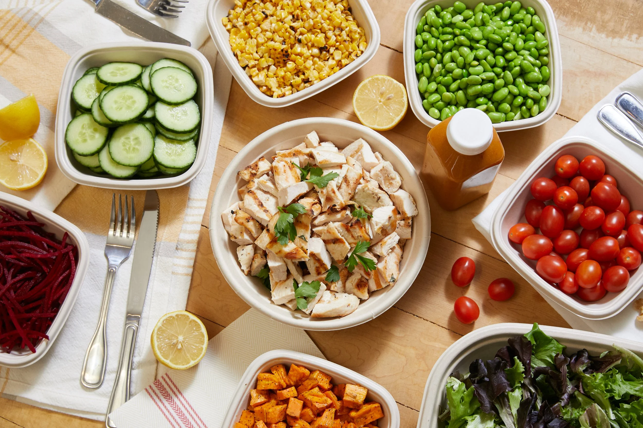 A spread of fresh vegetables and grilled chicken on a wooden table, including cherry tomatoes, cucumbers, corn, green peas, sweet potatoes, strawberries, and leafy greens, with lemon wedges and a bottle of salad dressing.