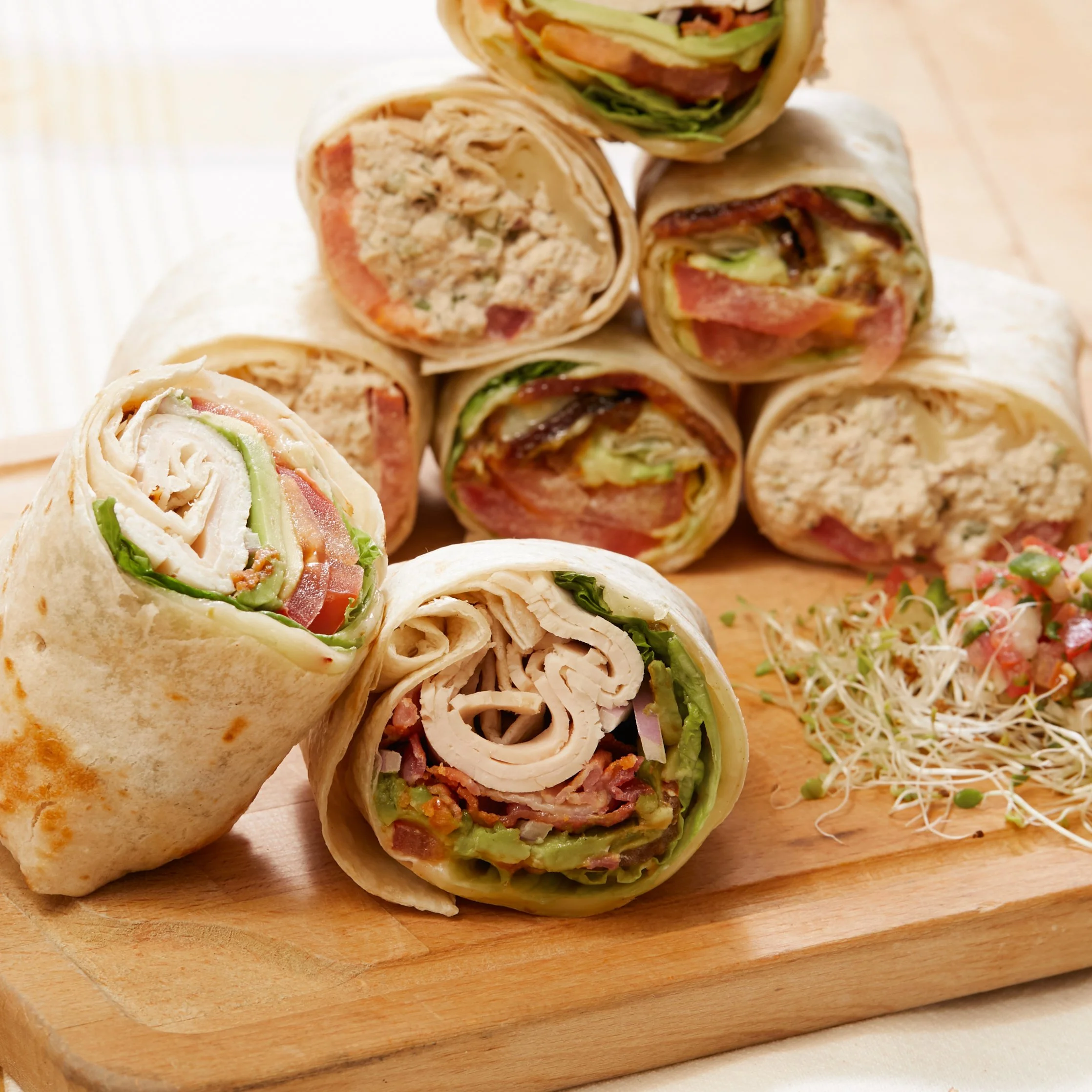 Assorted wrap sandwiches on a wooden cutting board with sprouts and chopped vegetables.