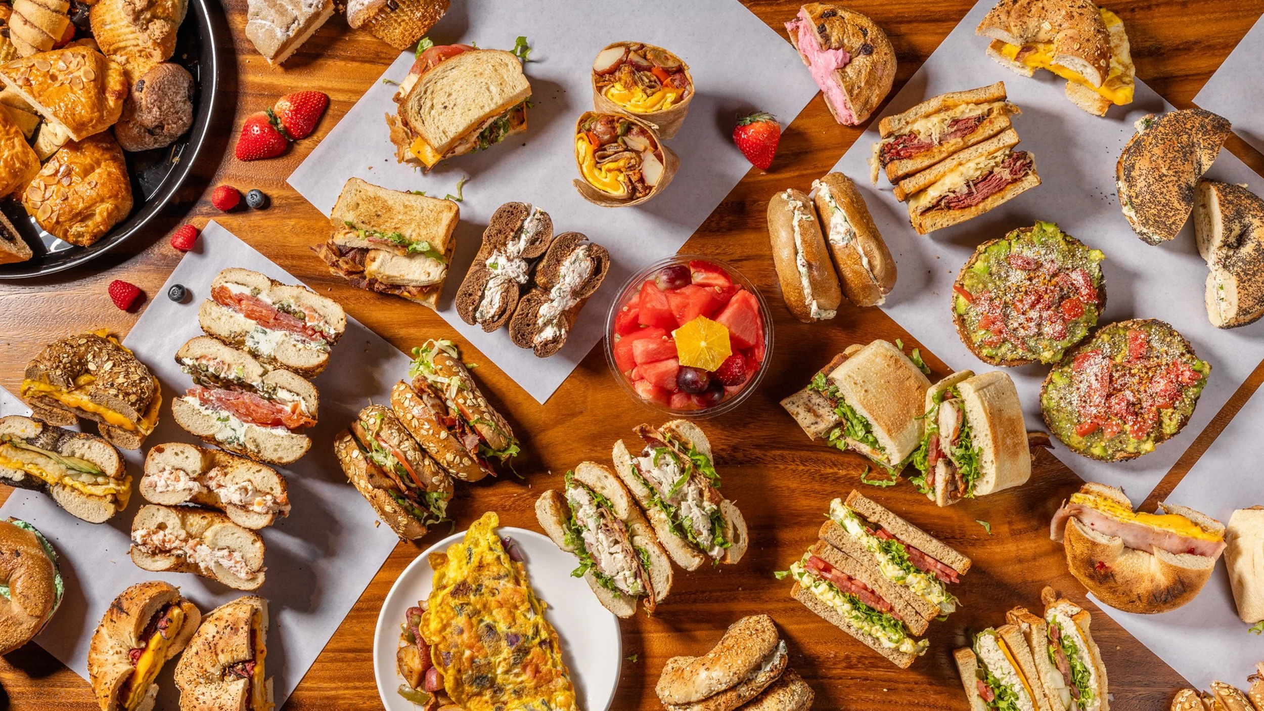 An overhead view of various sandwiches, pastries, and a bowl of fruit on a wooden table.