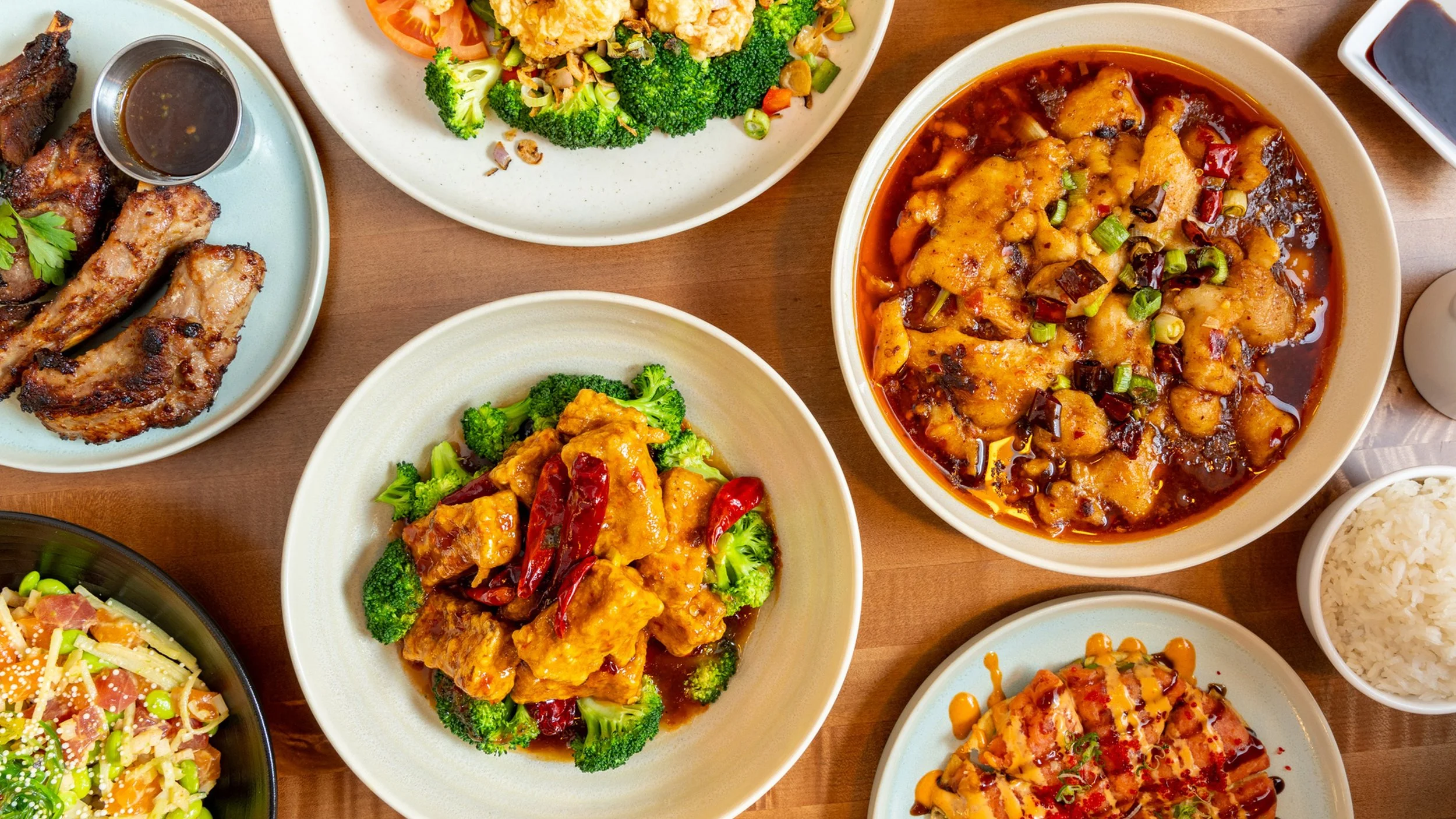 Assorted Asian dishes including chicken wings, broccoli with chicken in sauce, fried rice, and other flavorful stir-fried dishes on plates and bowls on a wooden table.