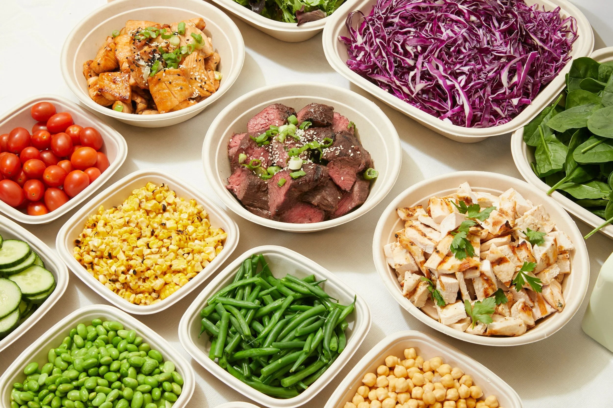 Bowl of cooked chicken with green onions, bowl of cooked red cabbage, bowl of fresh spinach, bowl of cherry tomatoes, bowl of roasted corn, bowl of cucumber slices, bowl of green beans, bowl of chickpeas, bowl of sliced cooked chicken, bowl of cooked