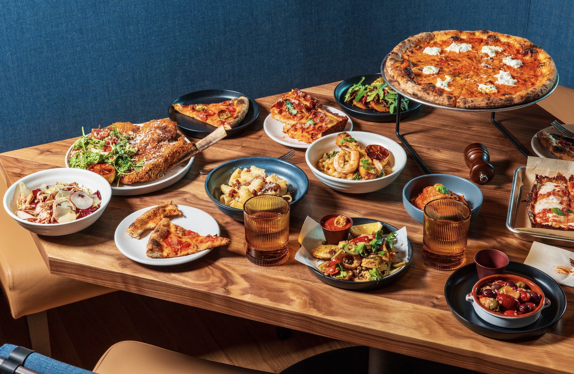 A wooden dining table filled with various dishes including pizza, salads, pasta, and bread, with glasses of iced tea and assorted plates of food.