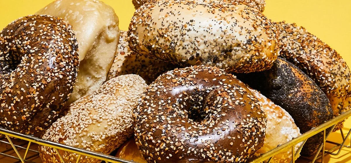 A basket filled with assorted bagels topped with sesame seeds, poppy seeds, and possibly onion or garlic flakes, set against a yellow background.