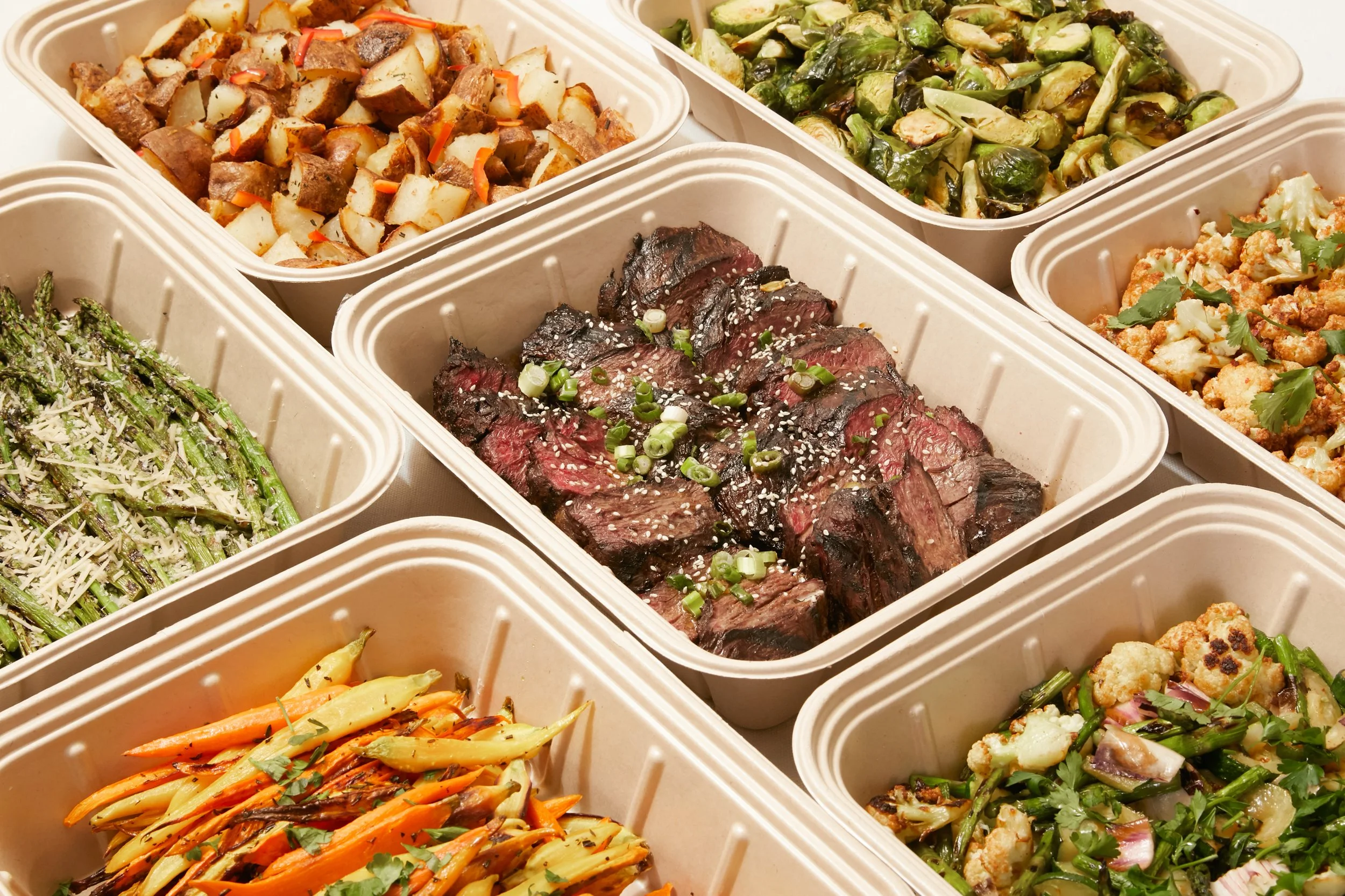 Assorted cooked food dishes in takeout containers, including grilled steak with green onions, roasted vegetables, roasted Brussels sprouts, roasted cauliflower, roasted carrots, and other vegetable dishes.