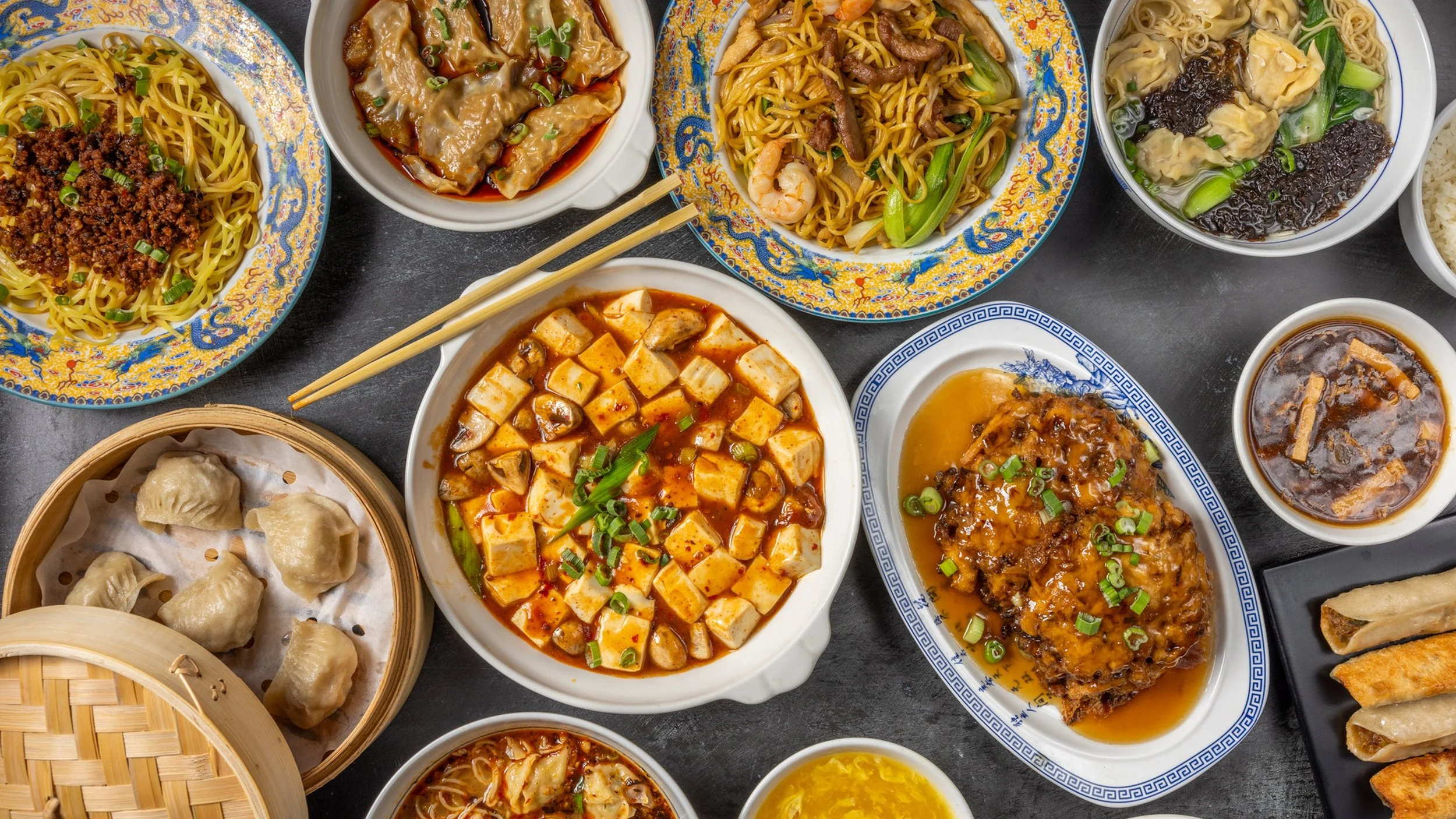 A variety of Chinese foods including steamed dumplings, Mapo tofu, noodle dishes with shrimp, beef, and vegetables, a bowl of soup with wontons, and sweet egg custard, all served on decorative plates and bowls.
