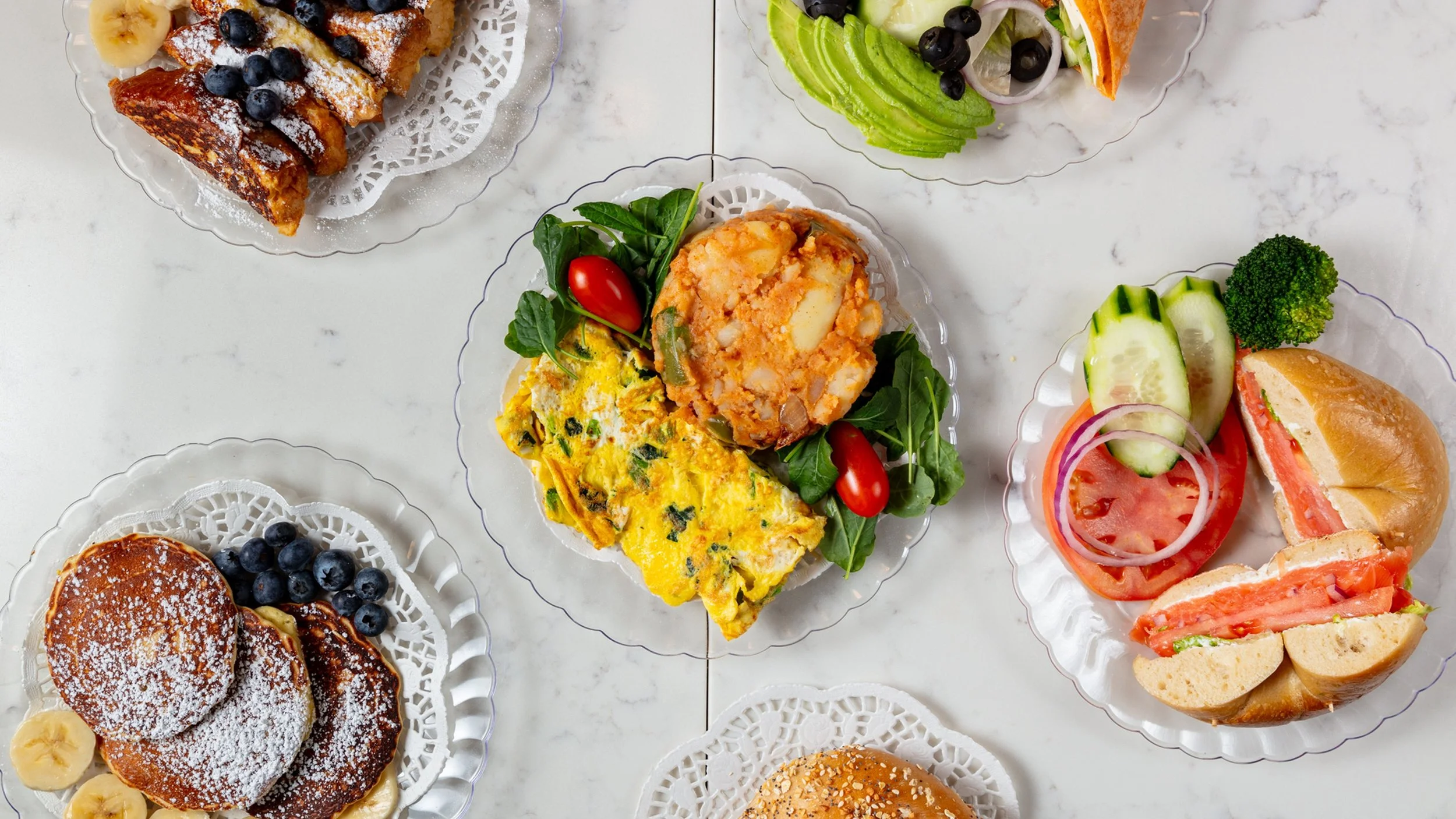 Assorted breakfast dishes including pancakes with blueberries and powdered sugar, an omelette with vegetables and cherry tomatoes, a veggie sandwich with cucumbers, tomatoes, onions, and lettuce, and a plate with crispy fried items, banana slices, and blueberries.