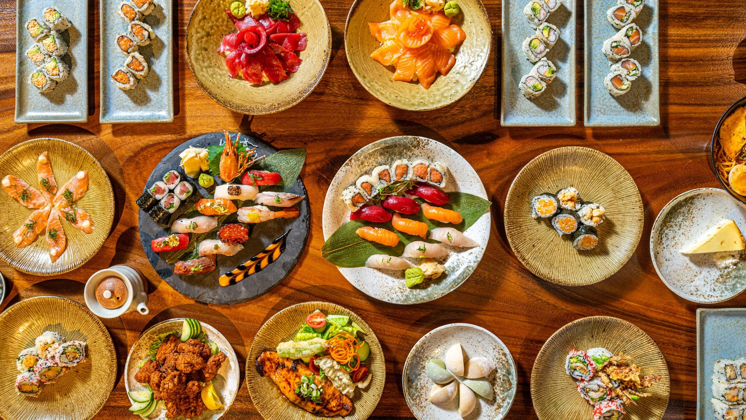 Assorted sushi dishes on various plates, including sashimi, nigiri, and rolls, arranged on a wooden table.