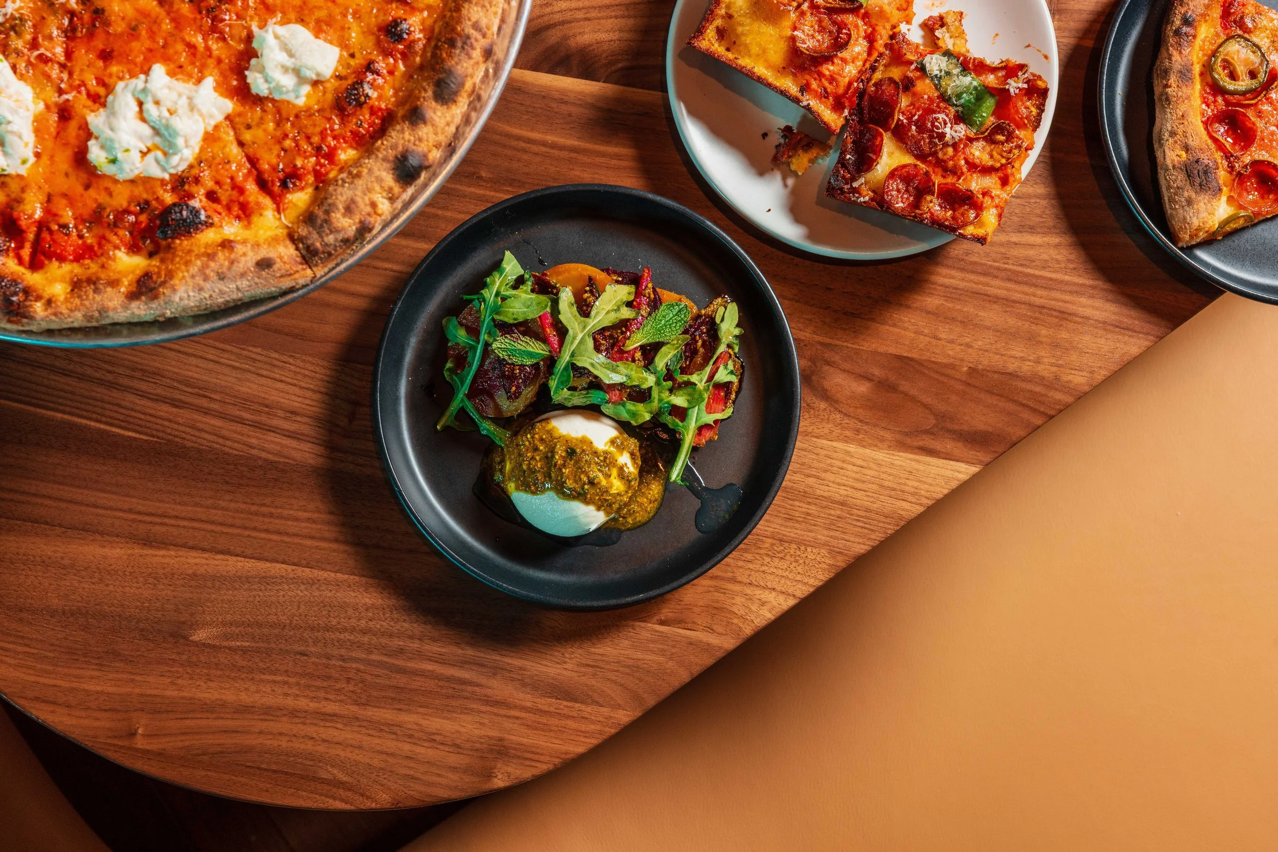 Assorted pizzas on plates and a salad with greens, vegetables, and a soft-boiled egg on a wooden table.