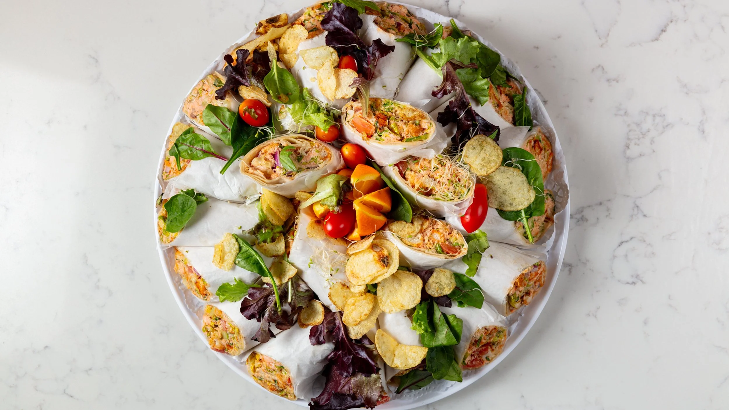 A plate of assorted spring rolls, cherry tomatoes, and vegetable chips on a white marble surface.