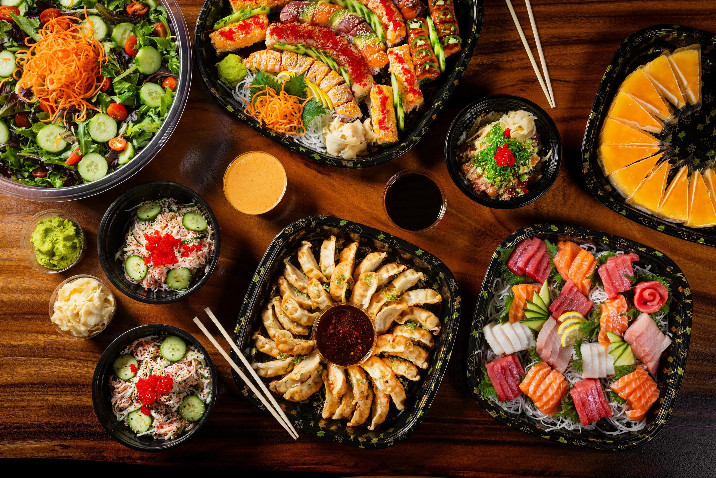 Assorted sushi dishes, sashimi, salad, and fruit platter on a wooden table.