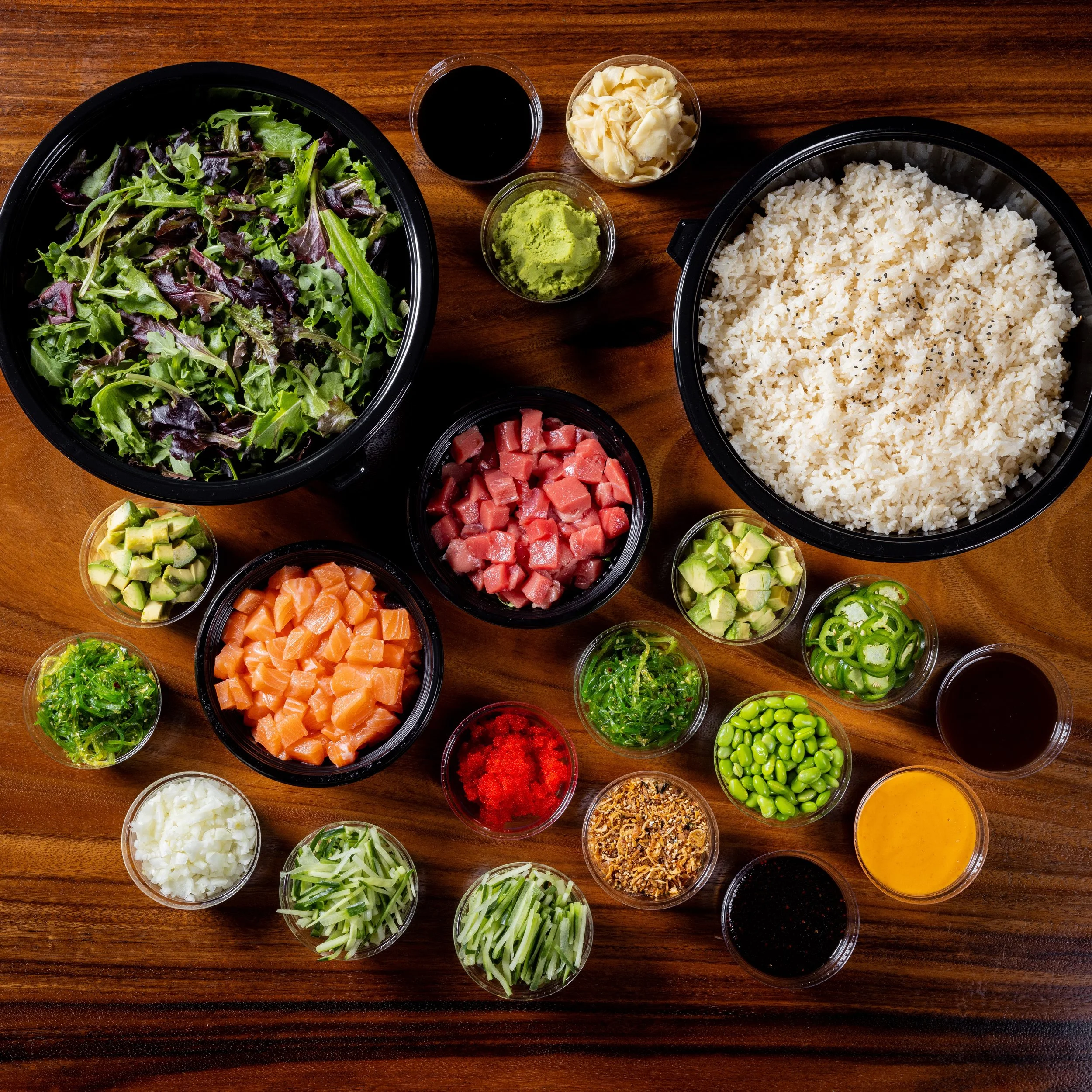 Assorted ingredients for making sushi, including rice, raw fish, vegetables, and sauces, arranged on a wooden surface.