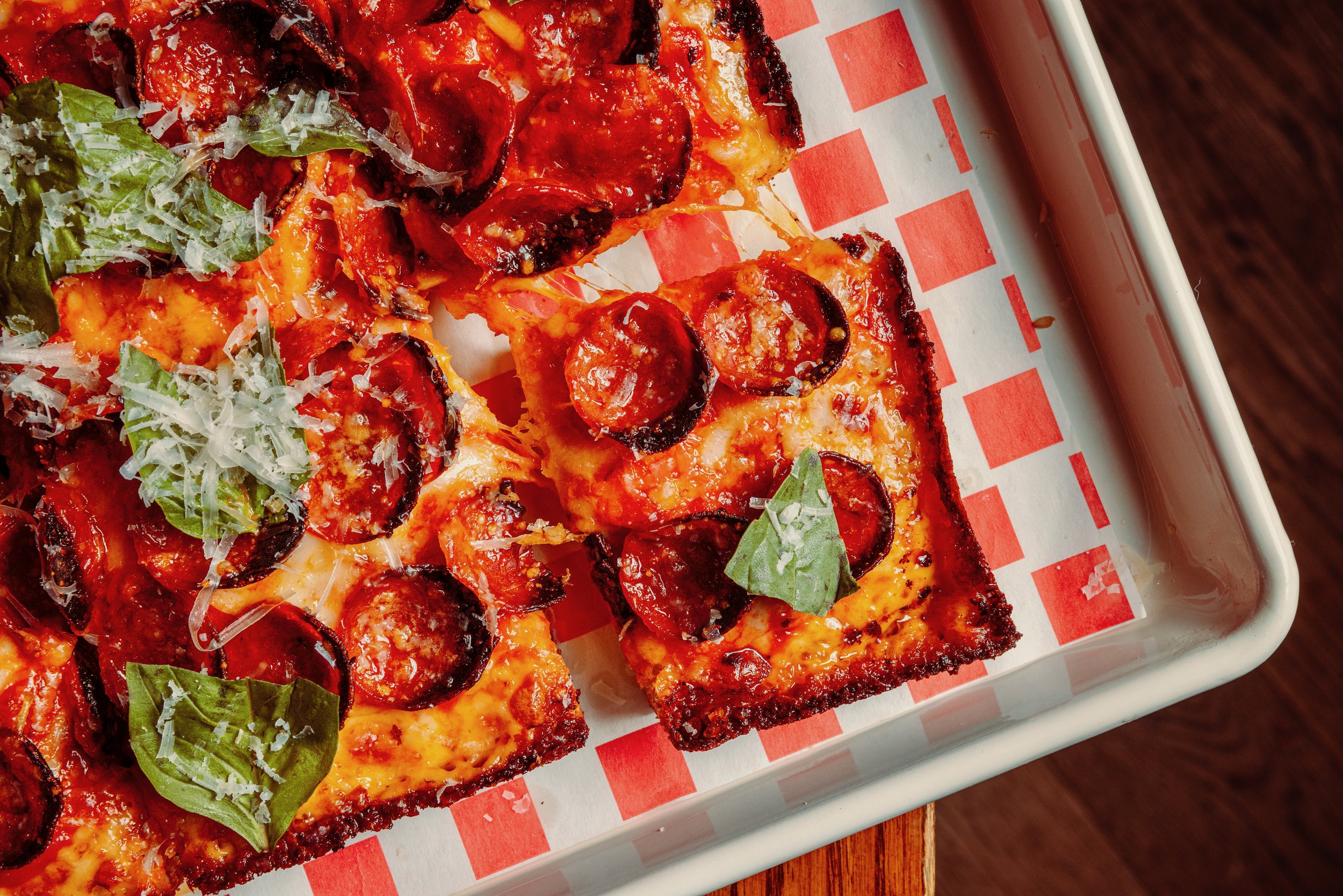 Close-up of slices of pepperoni pizza with melted cheese, fresh basil leaves, and grated cheese on a checkered paper-lined tray.