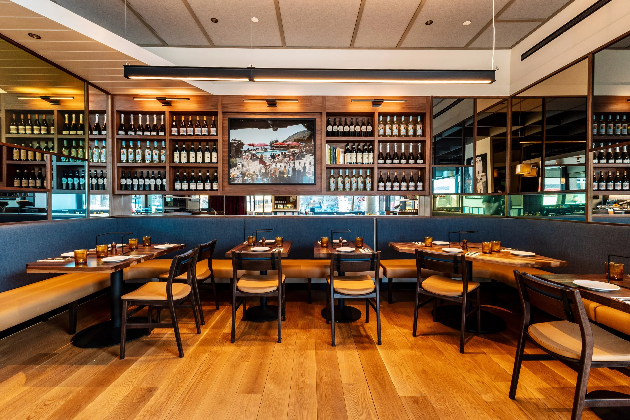 Interior of a modern restaurant with wooden floors, a curved booth with tables and chairs, and shelves of wine bottles on the walls.
