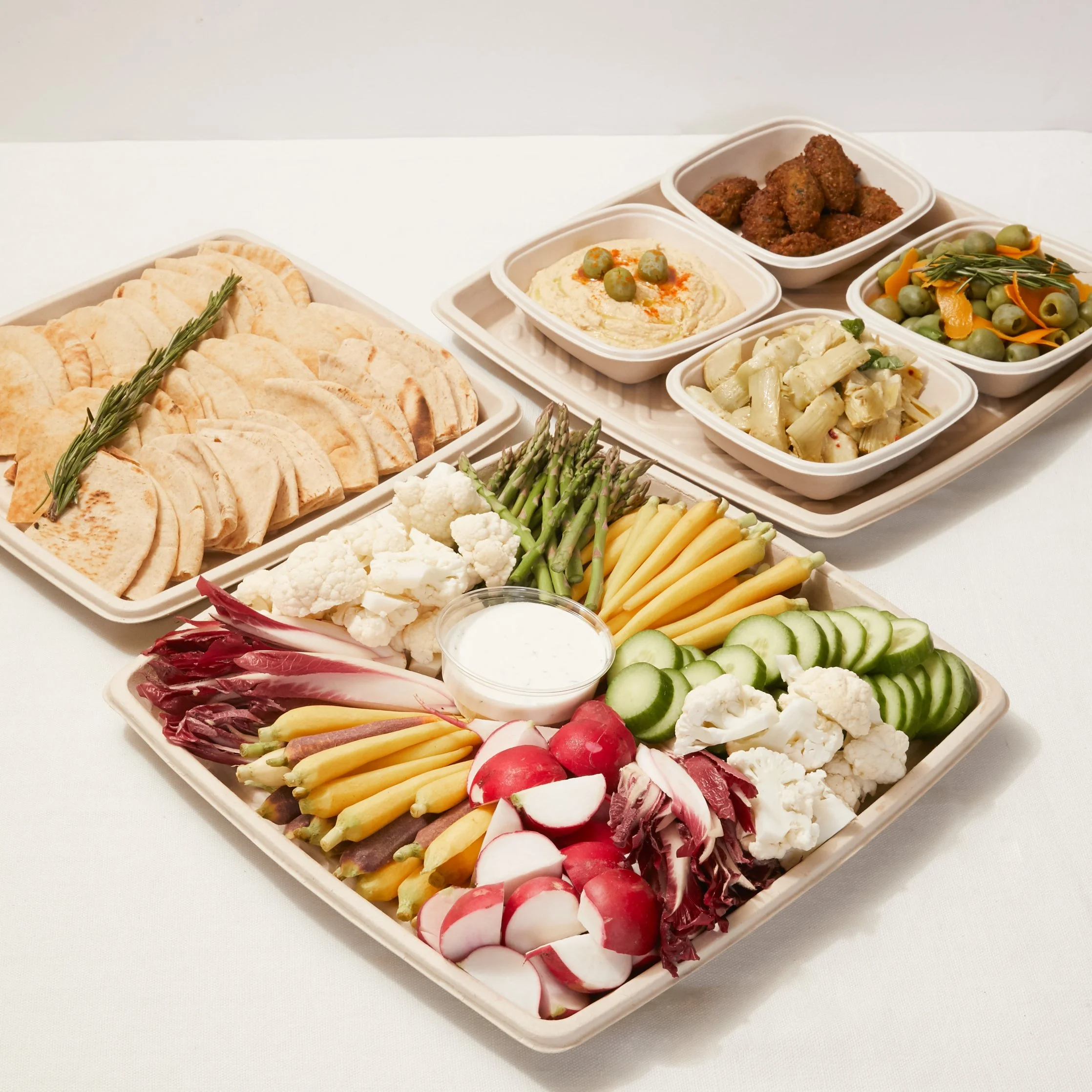 A large platter of fresh vegetables with radishes, cucumbers, cauliflower, asparagus, yellow and purple carrots, radicchio, and celery, with a container of dipping sauce, placed on a white table. Behind it, there are trays of pita bread, hummus, and 
