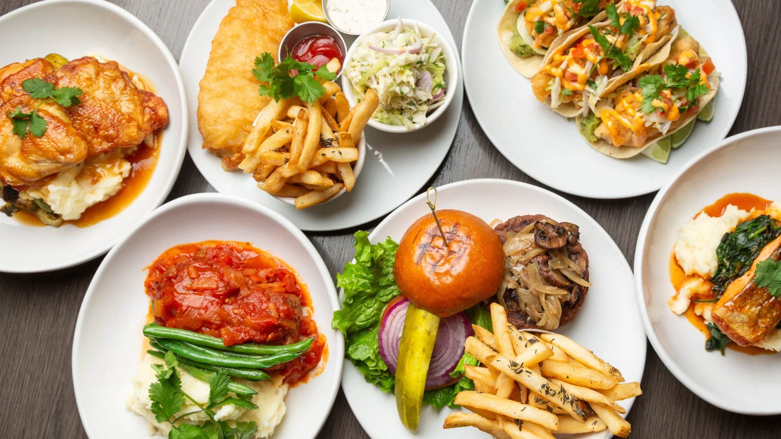 Assorted plates of food including chicken parmesan with mashed potatoes, fish fillet, French fries, coleslaw, tacos with cheese and cilantro, meatloaf with gravy, green beans, and a burger with lettuce, tomato, and onion.