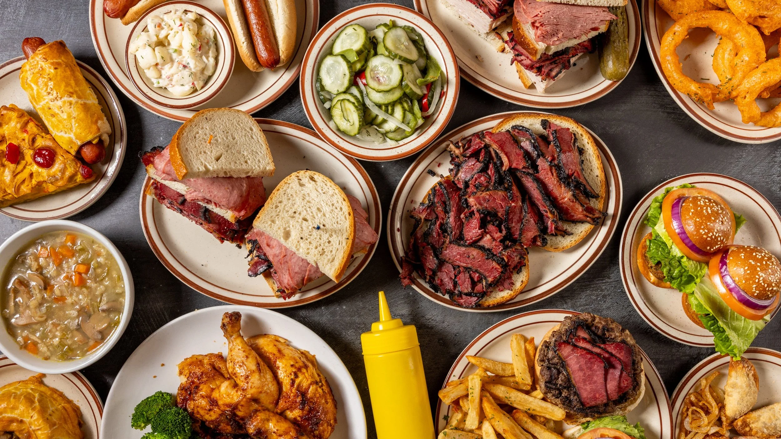 Assorted barbecue dishes and sides, including sliced beef brisket, hot dogs, onion rings, fried chicken, mini cheeseburgers, potato salad, cucumber salad, soup, and dipping sauce.