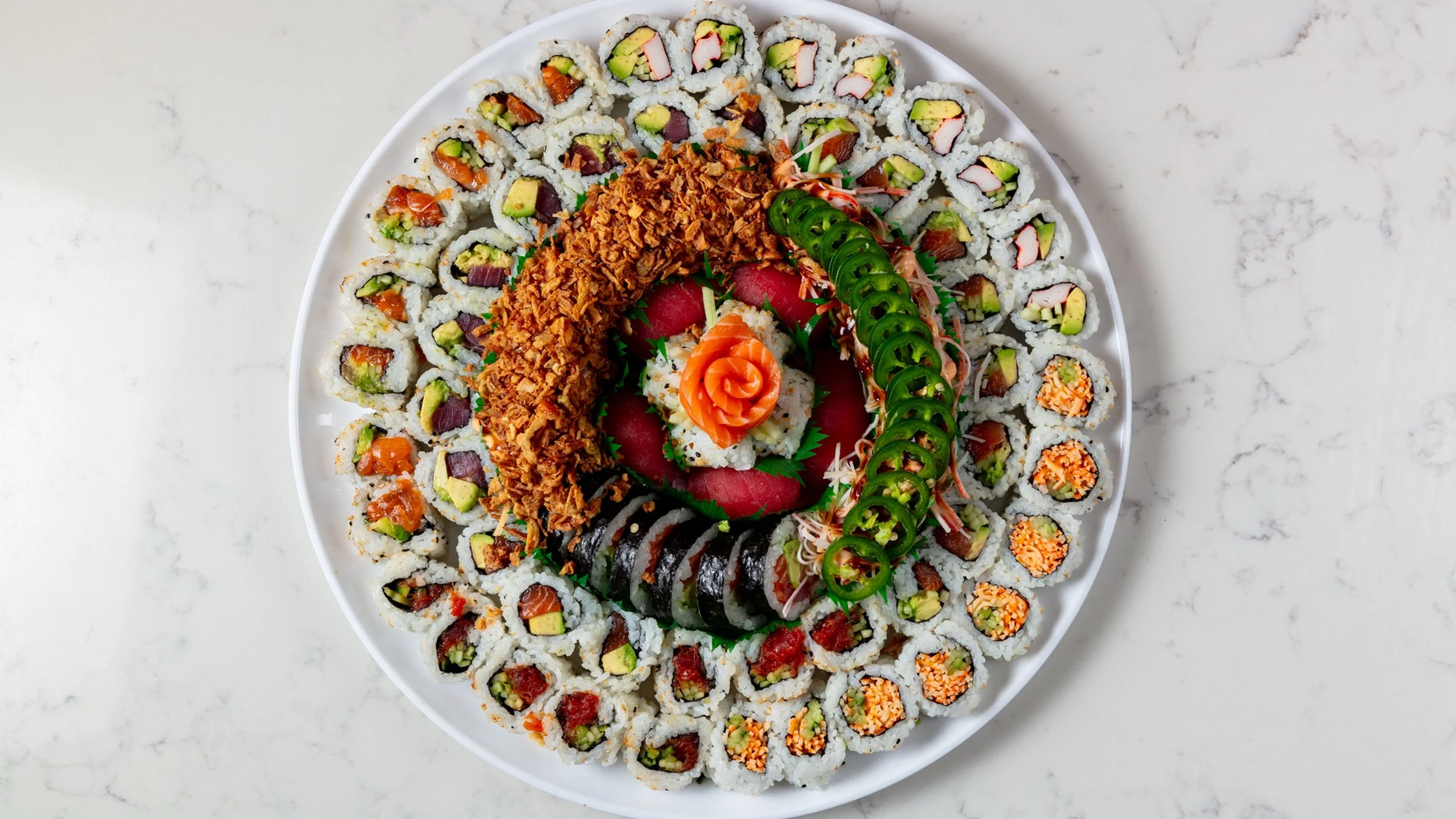 A large white plate filled with assorted sushi rolls, including California rolls, salmon, cucumber, and avocado, garnished with sliced jalapenos, shredded daikon, and a decorative salmon rose in the center, on a white marble surface.