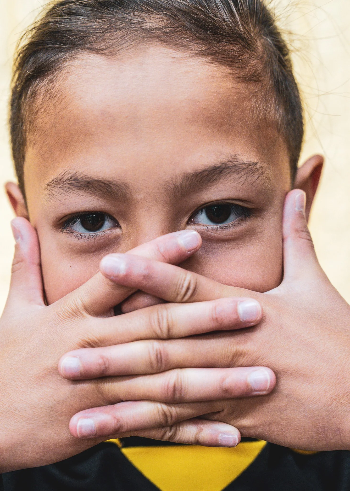 Close-up of a person with light skin covering their face with both hands, only eyes, forehead, and part of the nose visible, wearing a black shirt with a yellow design.