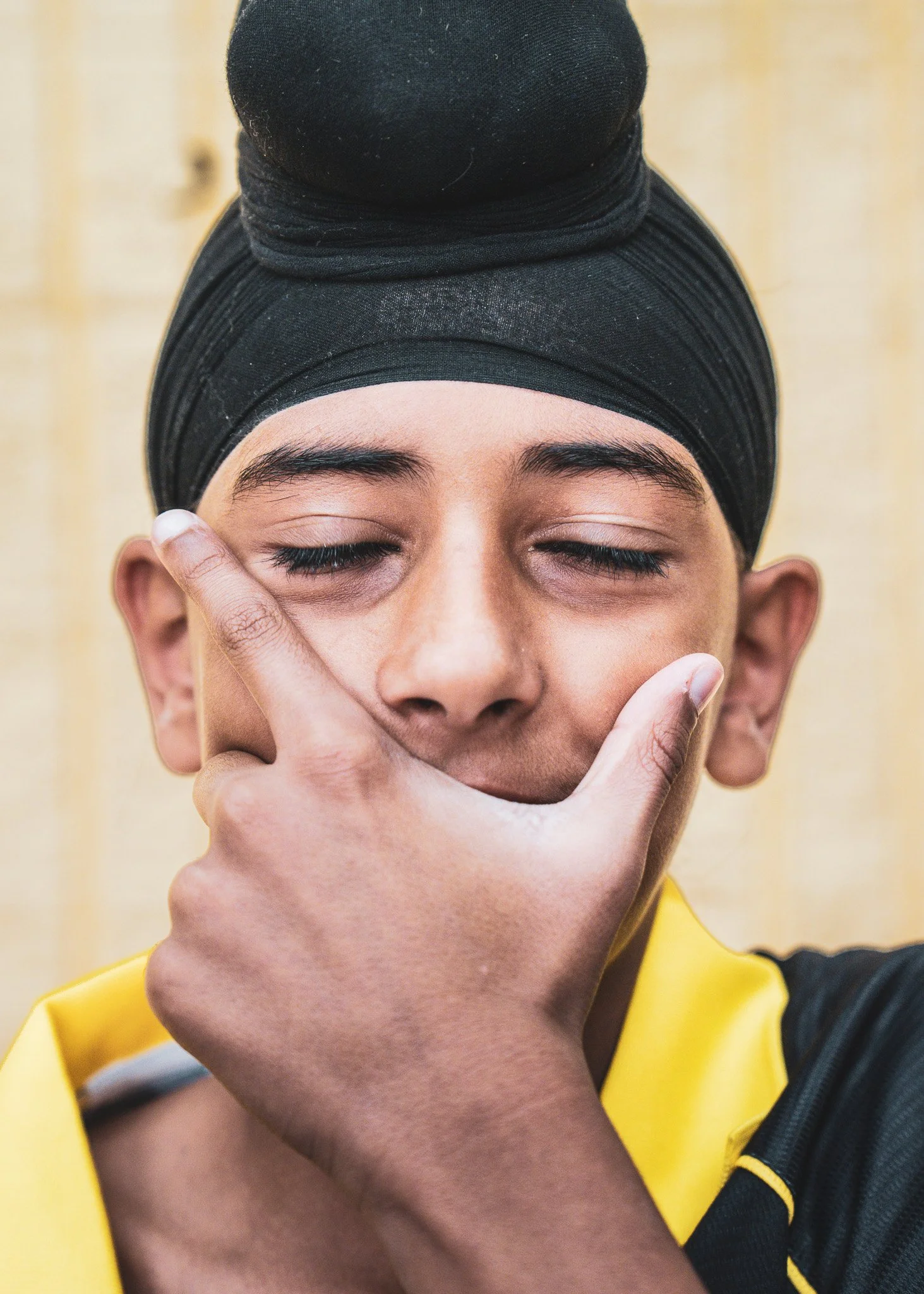 Close-up of a person with closed eyes wearing a black headscarf and a yellow and black sports shirt, touching their face near the temple with their right hand.