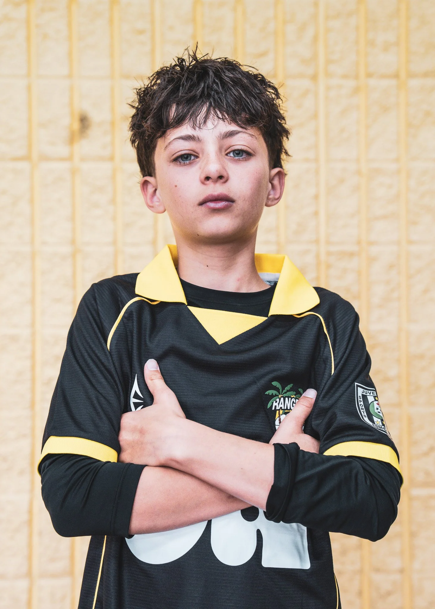 A person with short, curly dark hair, blue eyes, and light skin wearing a black sports jersey with yellow accents, standing against a yellow brick wall with his arms crossed.