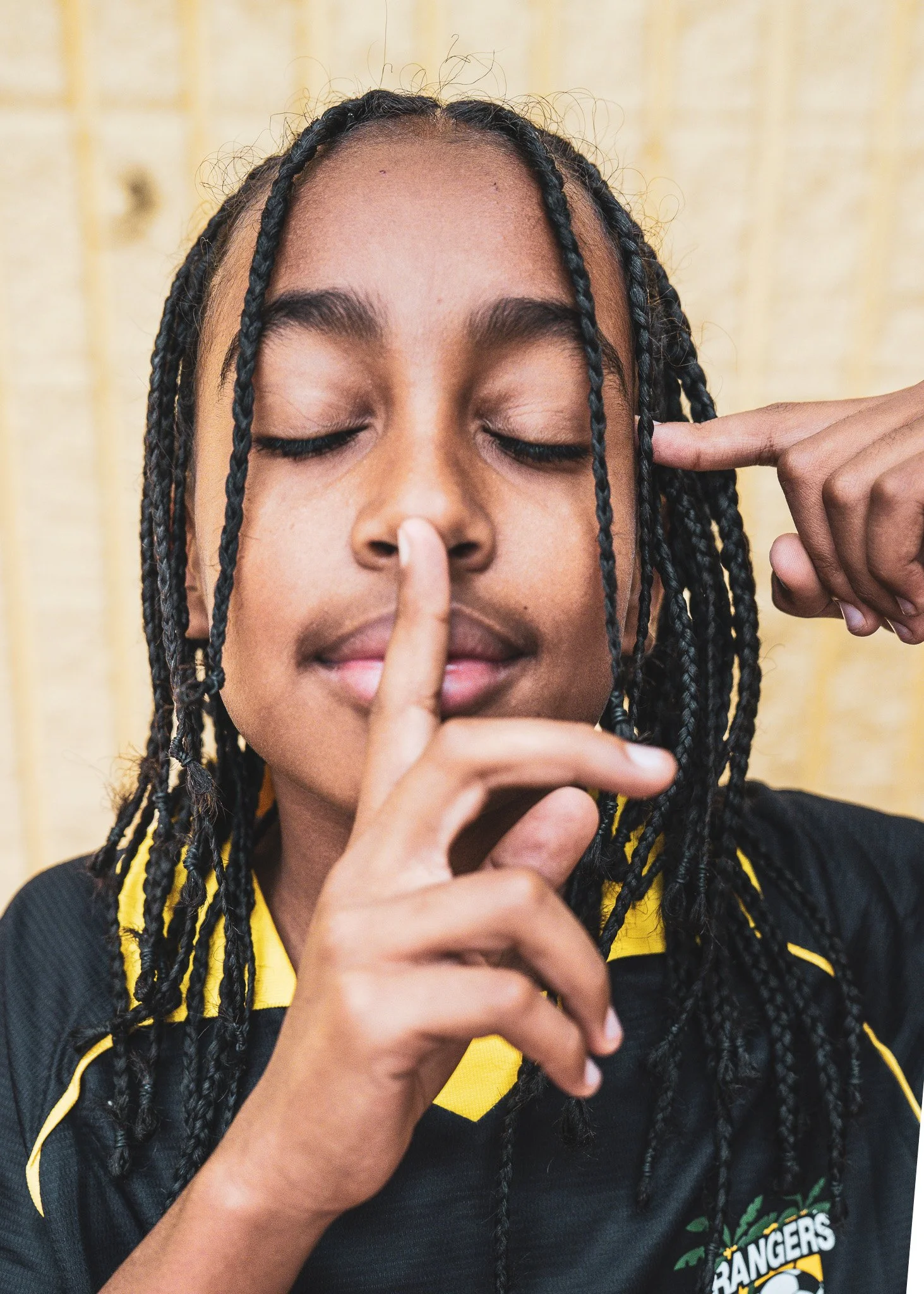 Close-up of a person with braided hair, eyes closed, holding index finger to her lips in a shushing gesture.