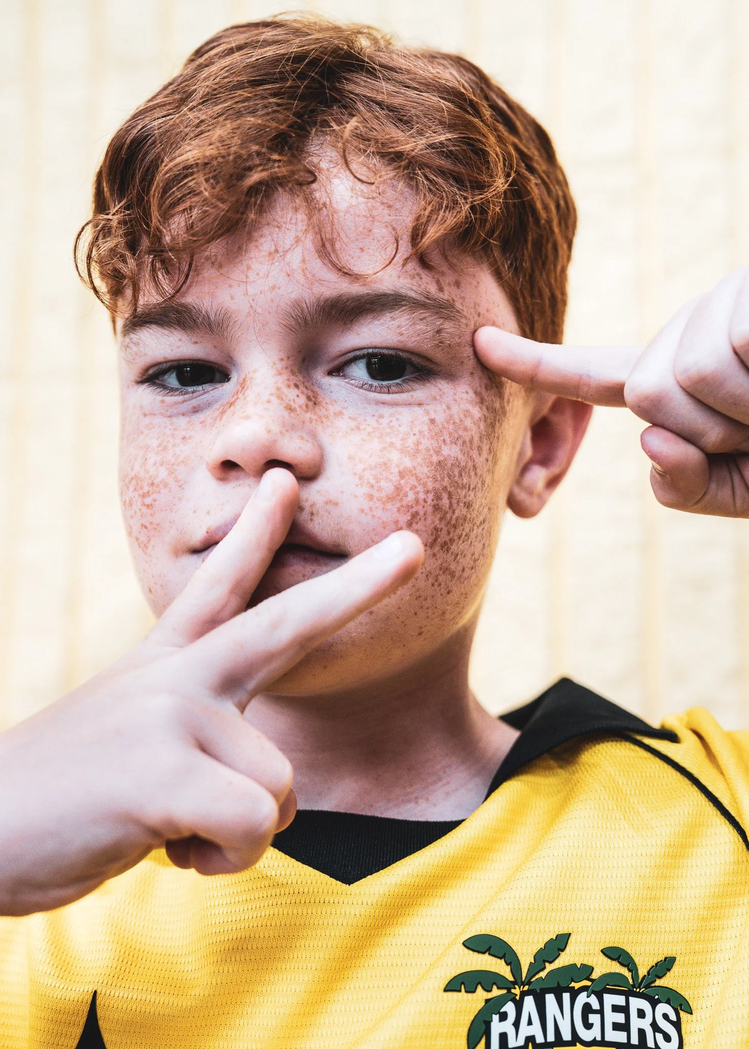 Close-up of a person with red hair and freckles, wearing a yellow sports jersey with black accents and a Halloween-themed patch with the word 'BANGERS' and a palm tree logo, making a finger gun gesture near his eye.