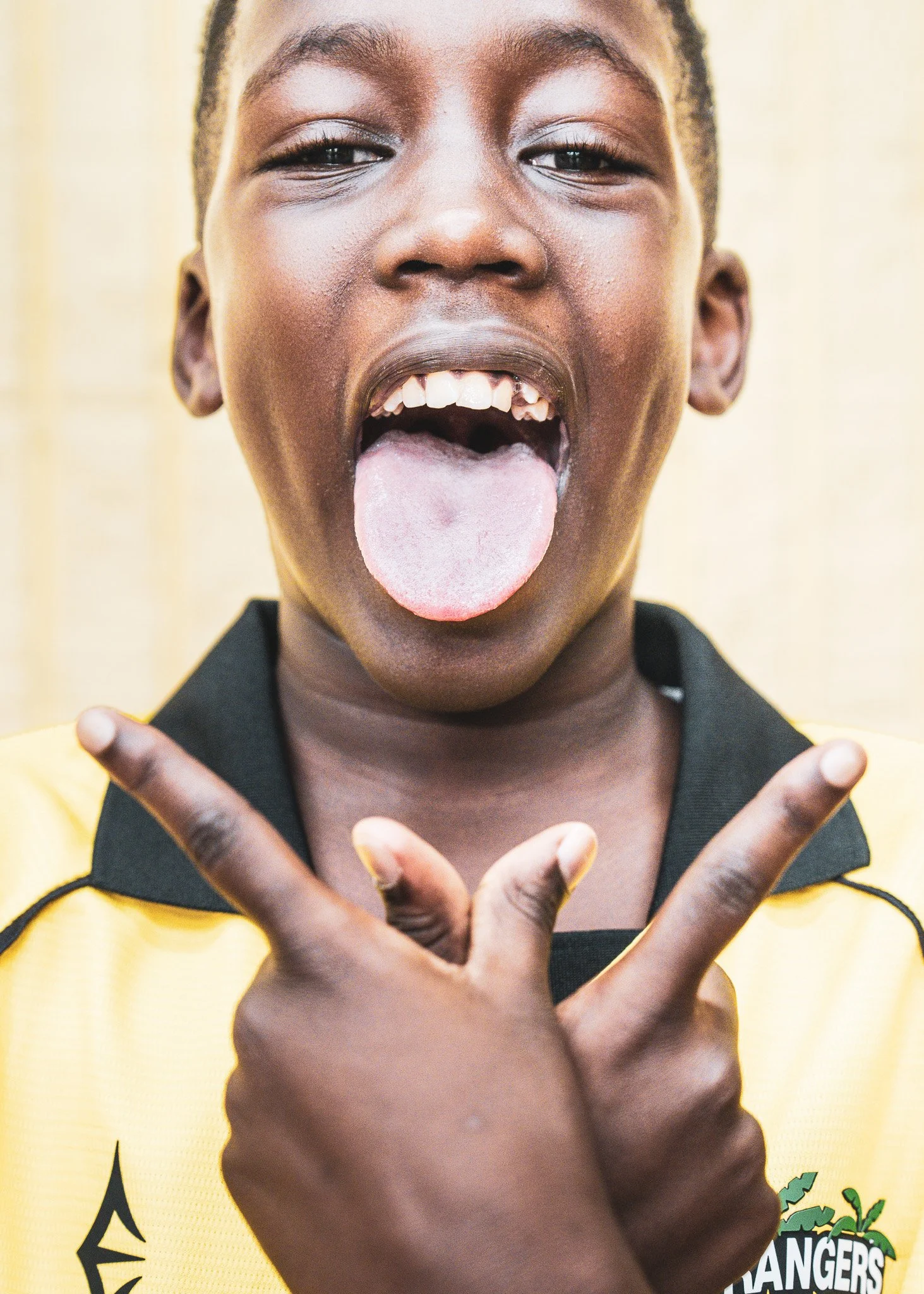 A person with dark skin sticking out their tongue and crossing their eyes, holding up their hands in a playful gesture, wearing a yellow and black shirt, with a blurred yellow background.
