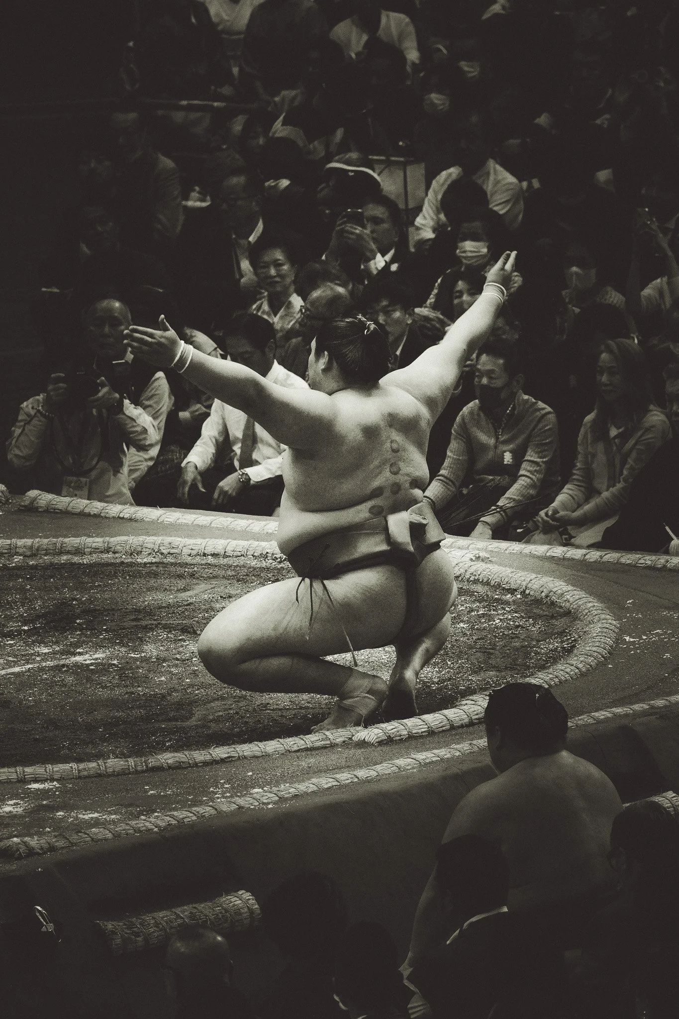 A sumo wrestler in a traditional event, squatting on a ring with one arm raised, surrounded by an audience in an indoor arena.