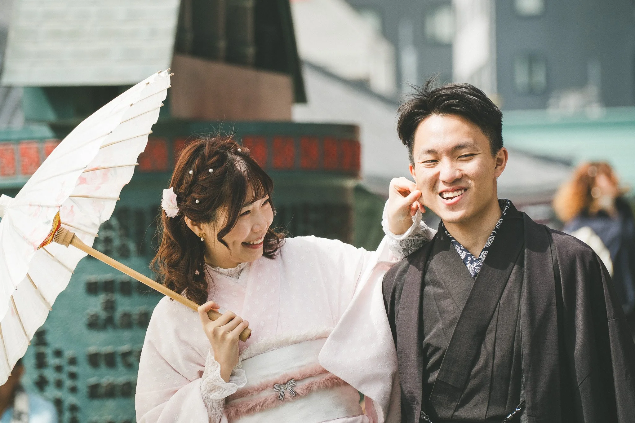 A smiling couple in traditional Japanese clothing, with the woman holding a parasol, against an outdoor urban backdrop.