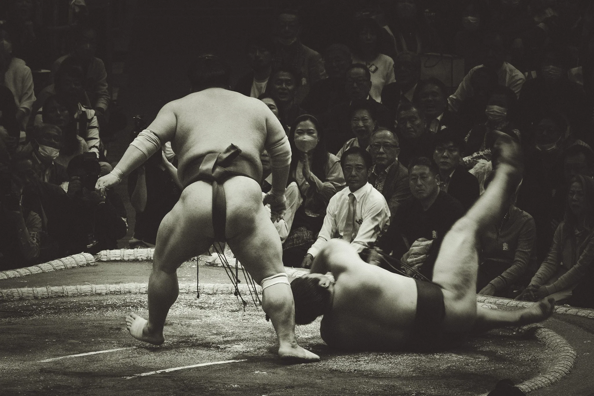 A sumo wrestler in traditional attire, facing away from the camera, in a sumo ring with a referee and an opponent on the ground, while an audience watches intently from the stands.