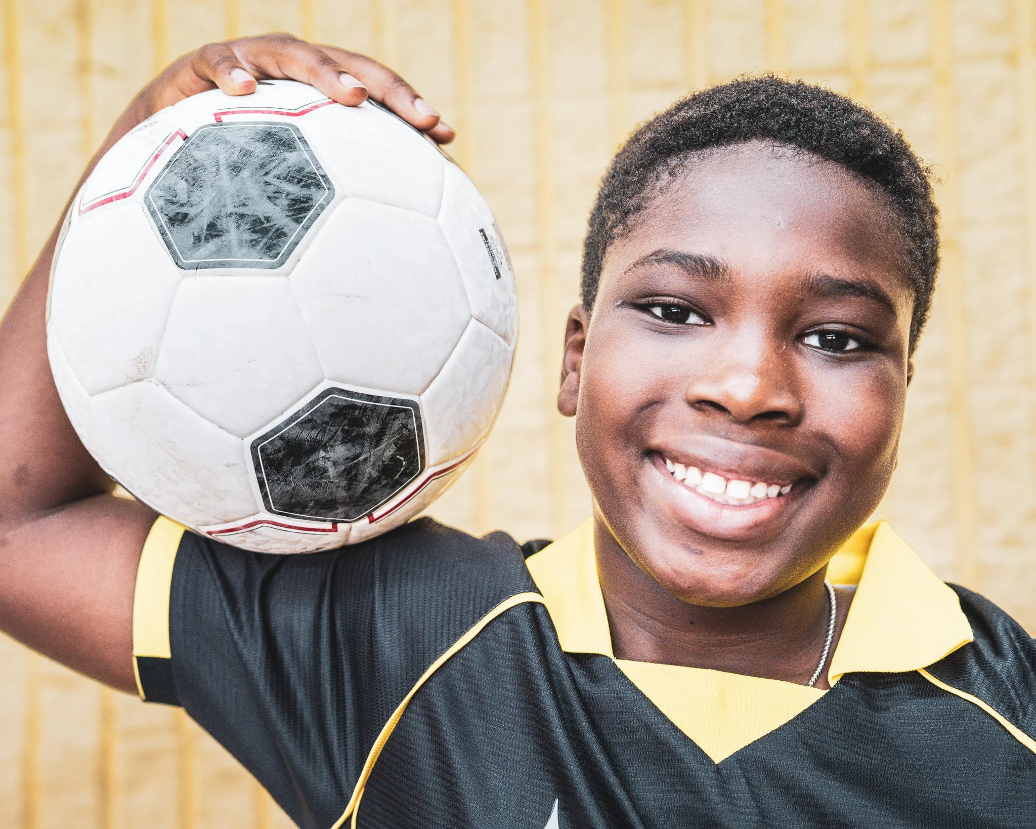 A person with a soccer ball over his shoulder, smiling, wearing a yellow and black sports jersey, with short curly hair, standing in front of a yellow wooden fence.