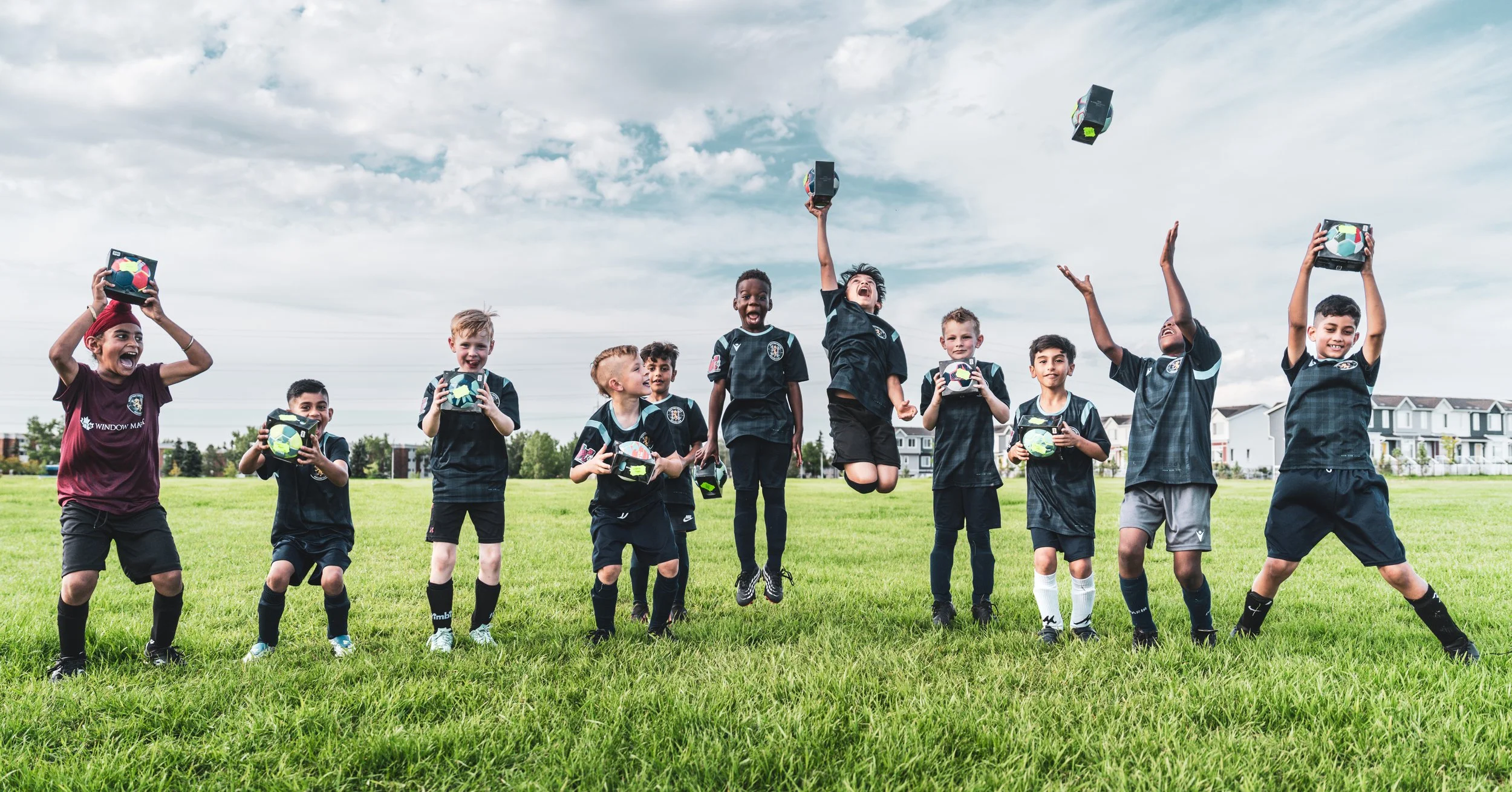 A team in soccer uniforms celebrating outdoors on a grassy field, holding colorful soccer ball lunchboxes, some jumping and some smiling, with houses and trees in the background under a partly cloudy sky.