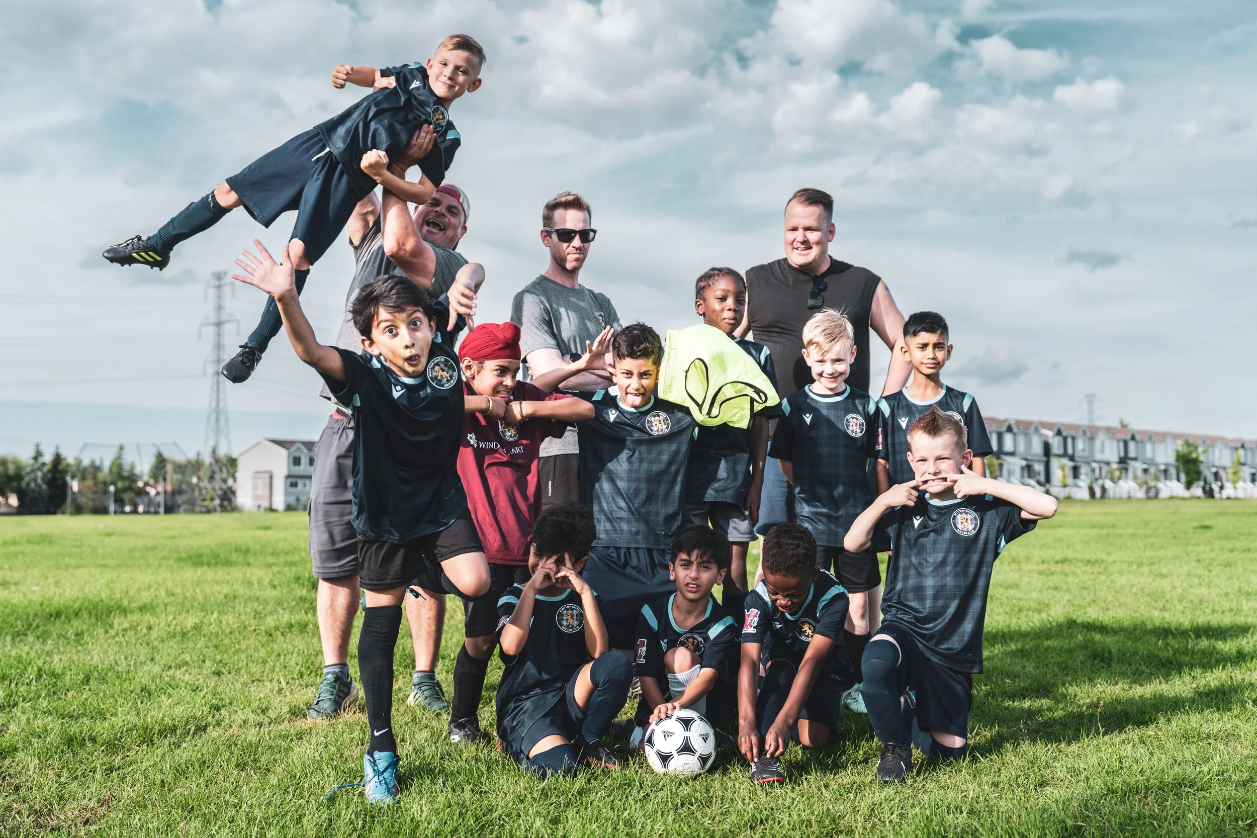 A group on a soccer field, posing for a team photo with a soccer ball in front. One child is being lifted and making a playful gesture, while others are smiling or making playful faces. The sky is partly cloudy, and there are houses and power lines i