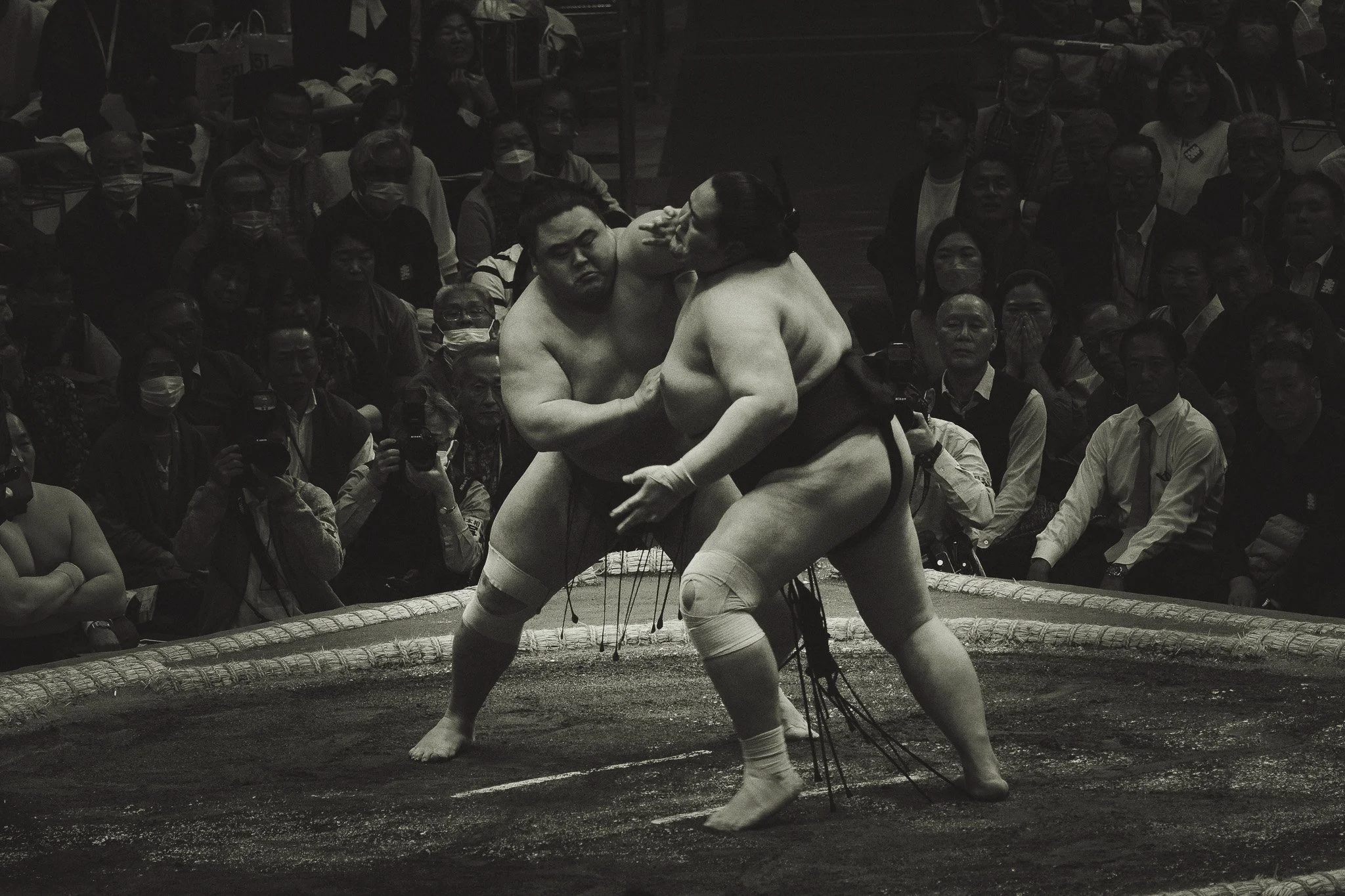 Two sumo wrestlers grappling in a ring surrounded by an audience, some wearing masks, during a sumo match.