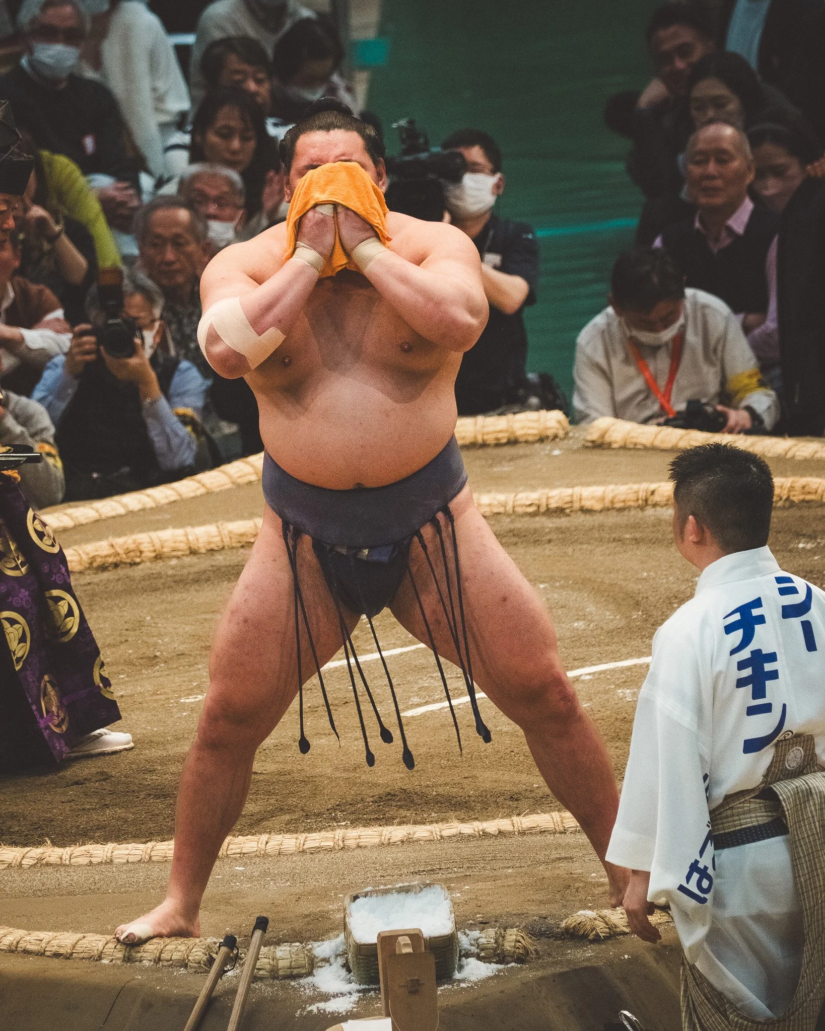 A sumo wrestler with a yellow towel over his forehead in a sumo ring, surrounded by spectators, officials, and photographers; soap and water are on the ground, indicating a ceremonial or cleaning ritual.