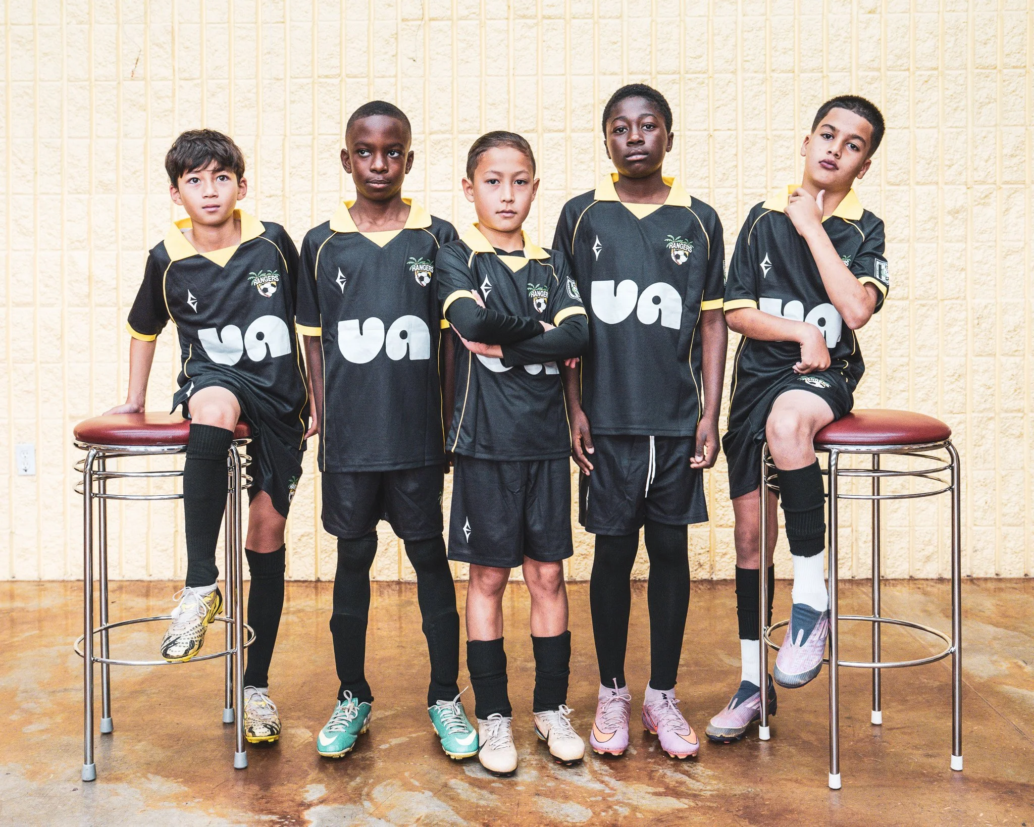 A group of five soccer players posing together indoors in black jerseys with yellow accents and a logo that says "Rangers." They are standing against a beige wall with vertical lines, with two of them sitting on stools and three standing behind them.