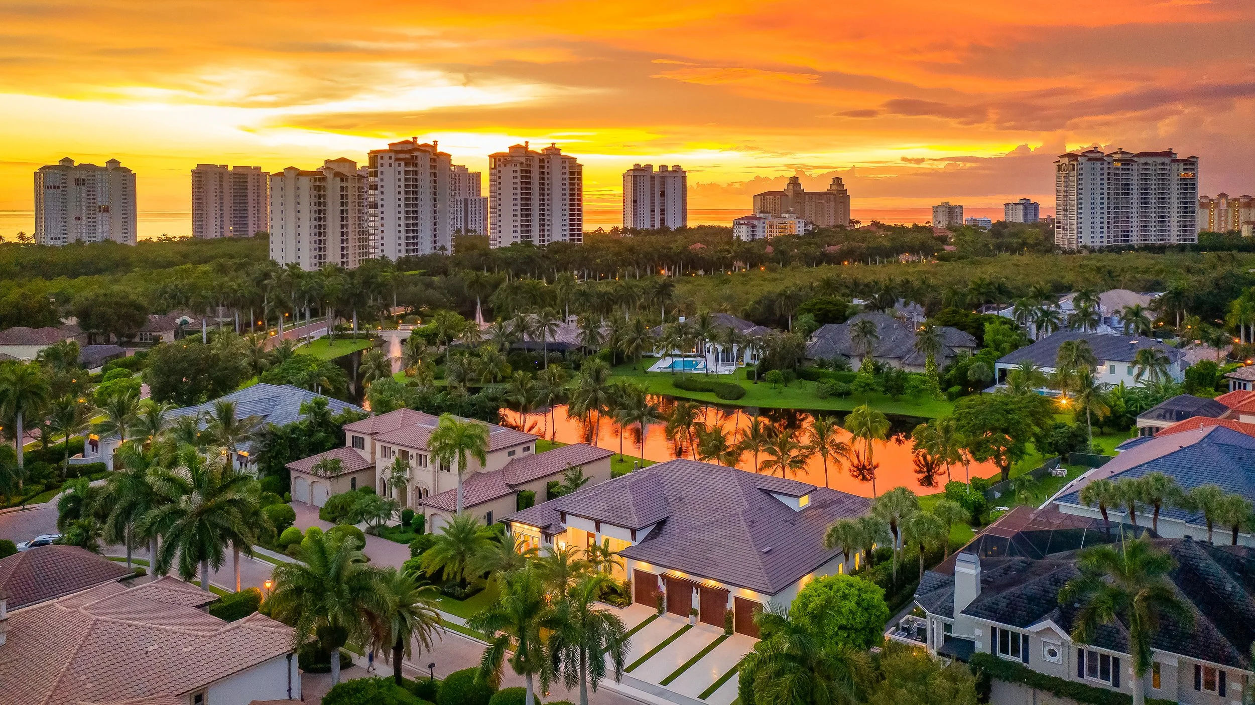Aerial view of a residential area with large houses, palm trees, a pond reflecting the sunset, and high-rise buildings in the background during sunset.