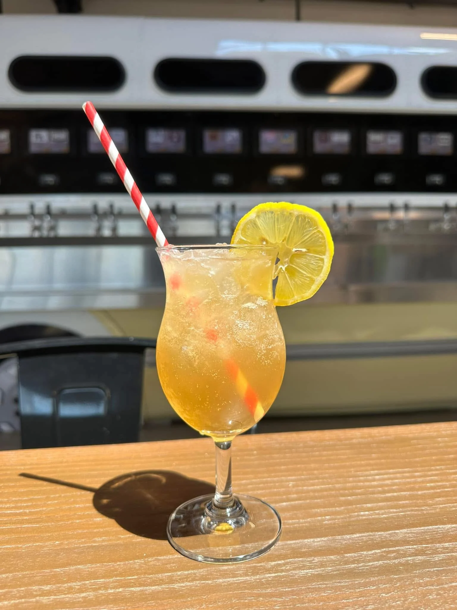 A tall glass of citrus cocktail garnished with a lemon slice and a red-and-white striped straw on a wooden table, with a stainless steel appliance in the background.