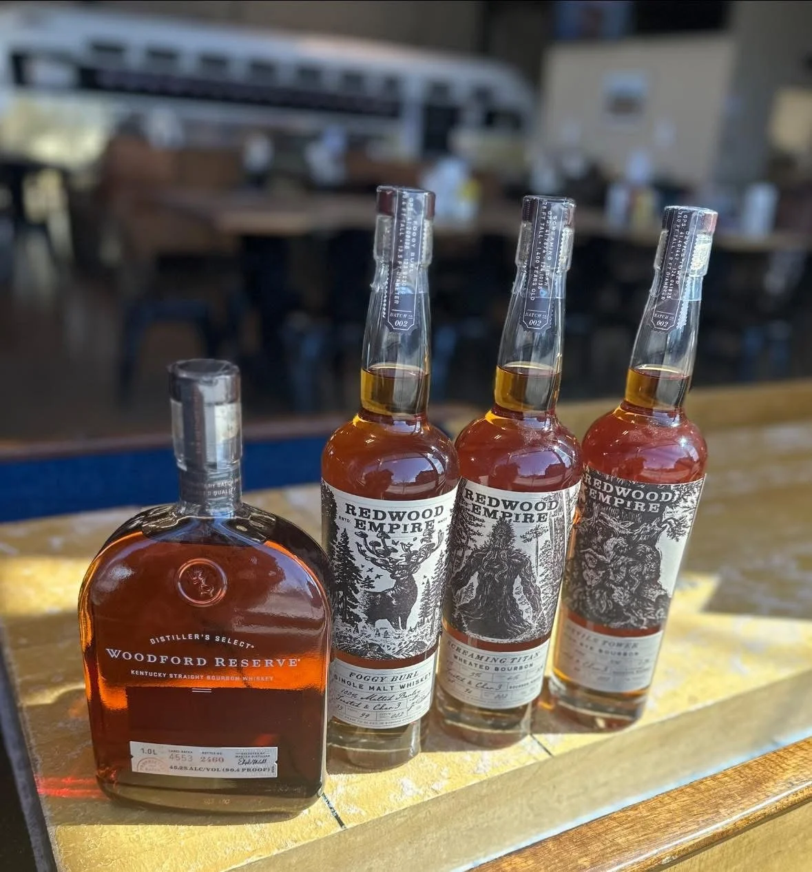 Four bottles of bourbon whiskey are displayed on a wooden surface in a bar setting. The first bottle is Woodford Reserve, and the remaining three are Redwood Empire, each with distinct labels featuring nature-themed illustrations.