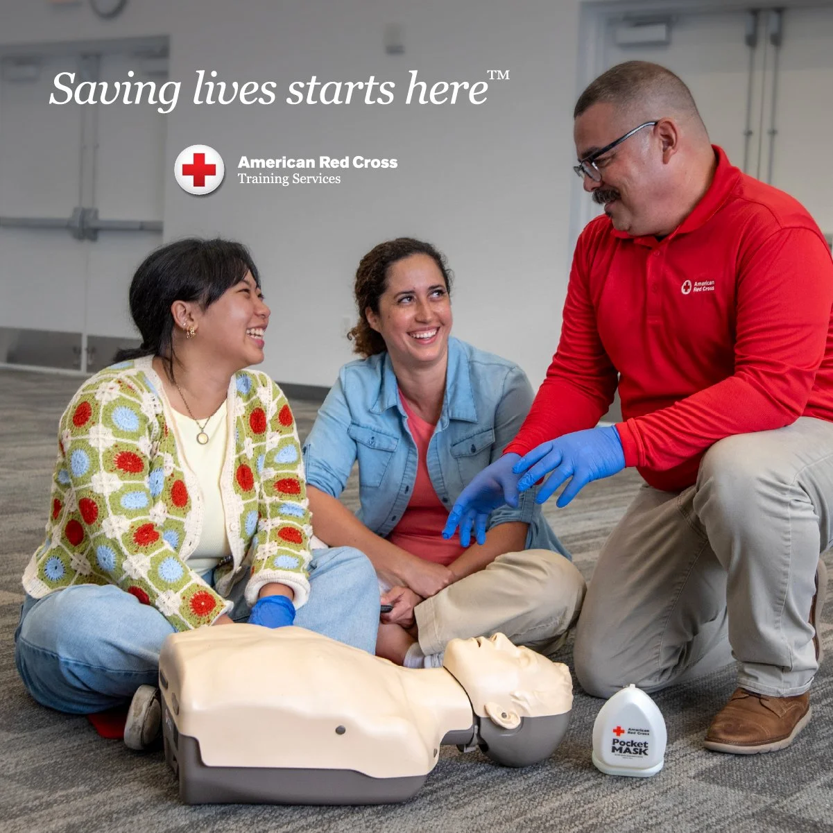 Three women participating in a CPR training session with an instructor demonstrating on a mannequin. They are sitting on the floor with training equipment around them, including a mask and a training dummy.