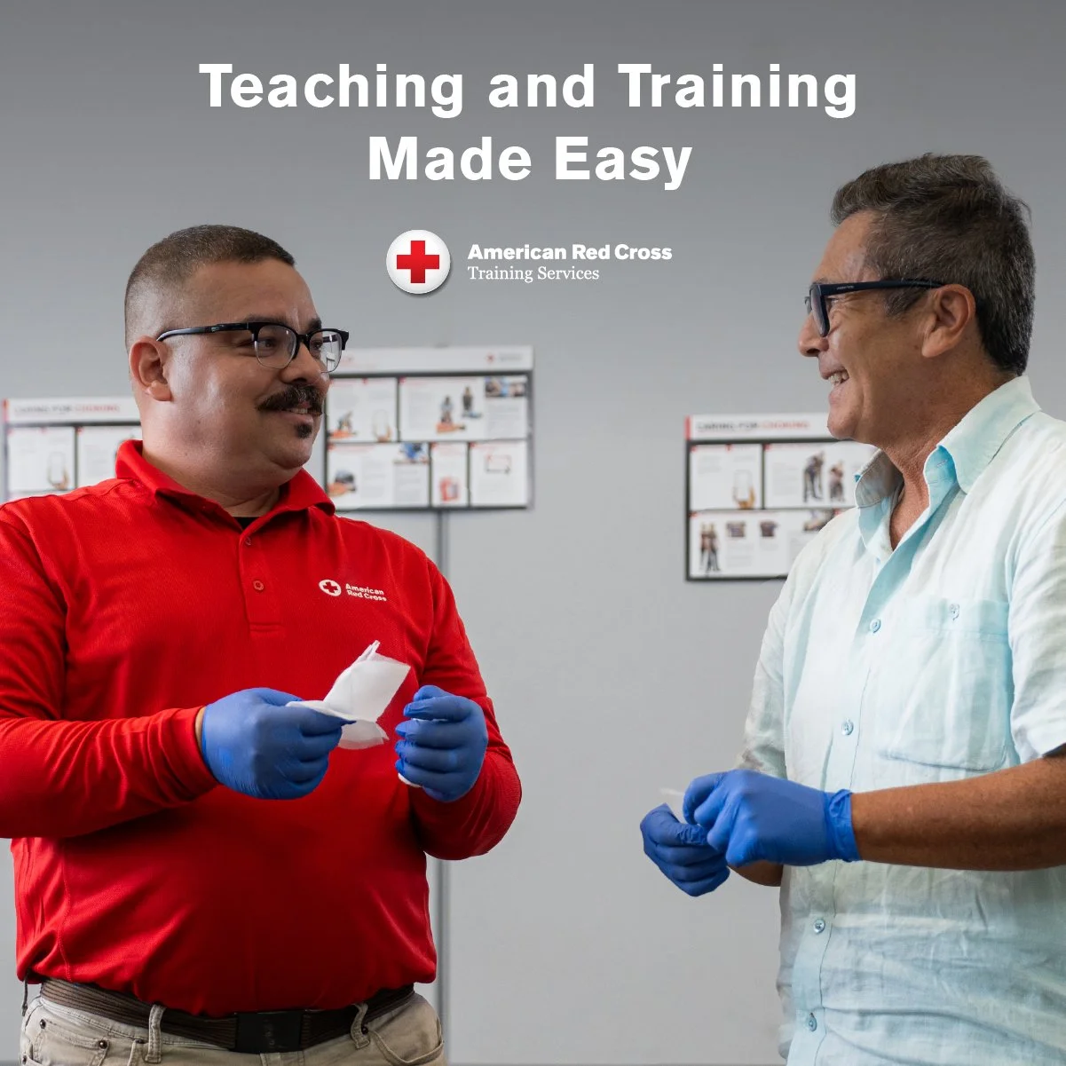 Two men in a training room, one wearing a red shirt with the American Red Cross logo, and the other wearing a light blue shirt, both wearing blue gloves and holding tissues, engaged in a conversation.