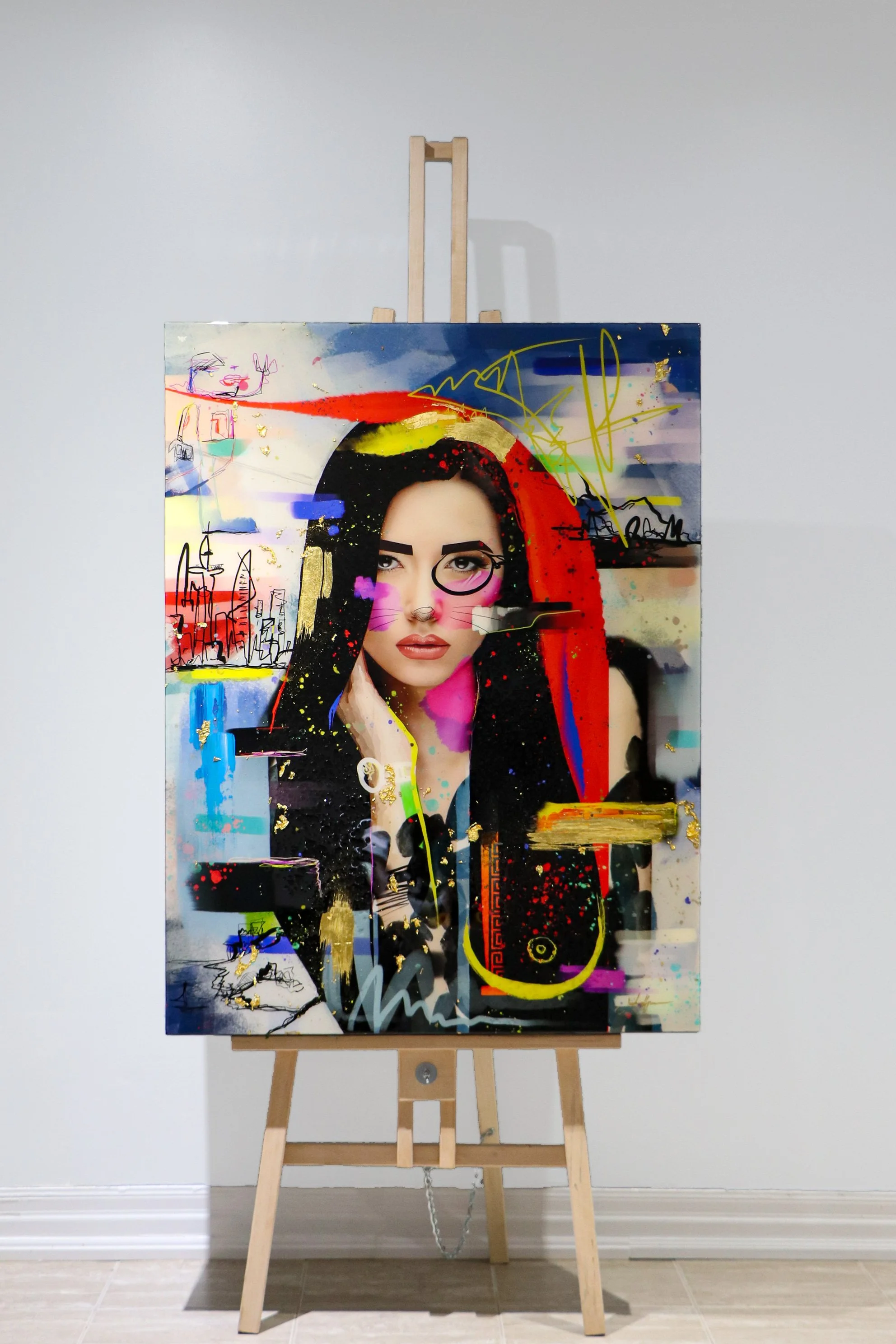 An abstract mixed-media portrait of a woman on canvas, featuring a realistic face with dark hair, colorful paint splashes, scribbles, and urban skyline sketches, displayed on a wooden easel.