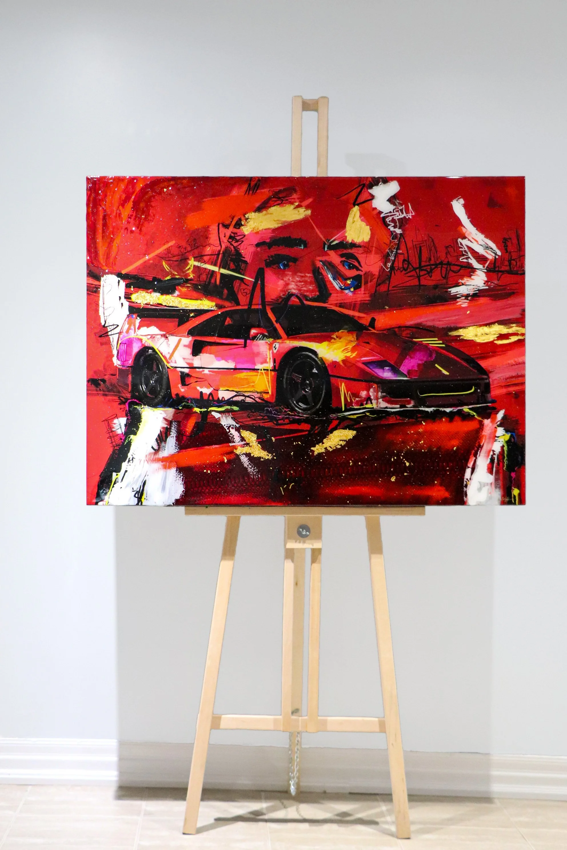 Abstract painting on a canvas displayed on a wooden easel, featuring a red sports car and a large face in a mix of vibrant colors and expressive brushstrokes.
